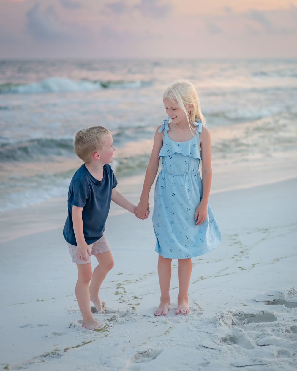 Two young blonde siblings holding hands and smiling on a sandy beach at sunset.