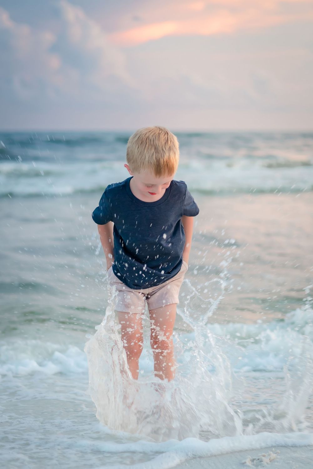 Young blonde boy playing in ocean waves on the beach, getting splashed by the water at sunset.