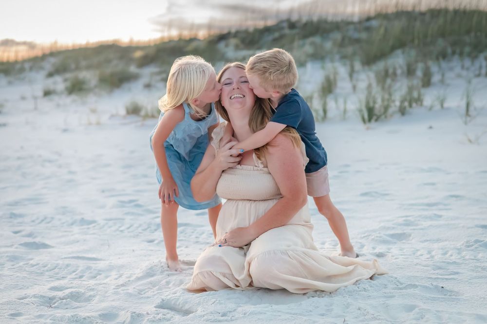 Pregnant mother sits on beach sand while her two young children lovingly kiss her cheeks.