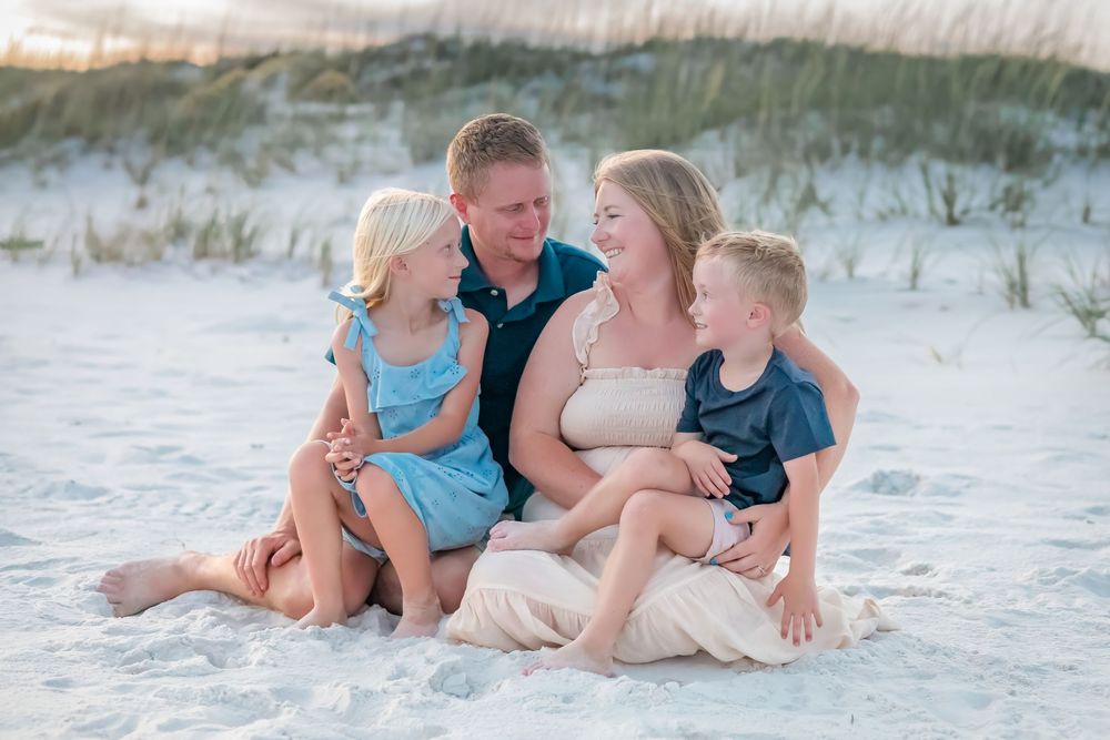 Laughing family of four sits together on white sand beach dunes at golden sunset.