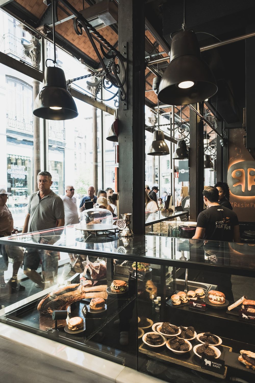 Busy gourmet market food stall with glass display case of pastries and desserts, industrial pendant lighting.