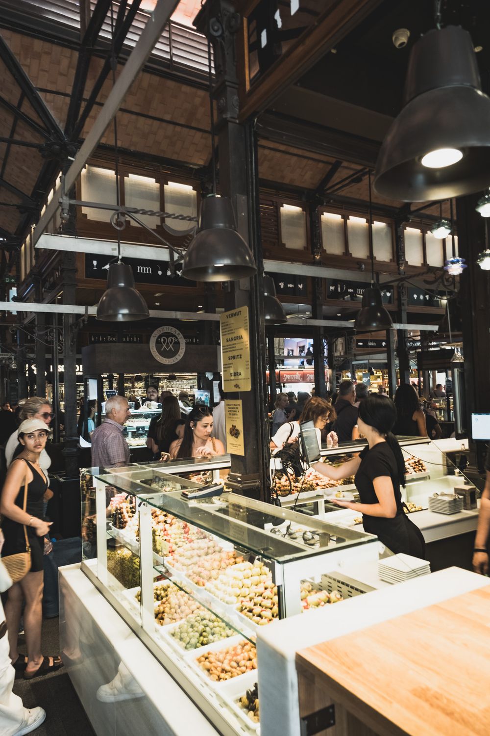 Bustling indoor gourmet market with food display counters, shoppers browsing stalls under high iron-framed ceiling.