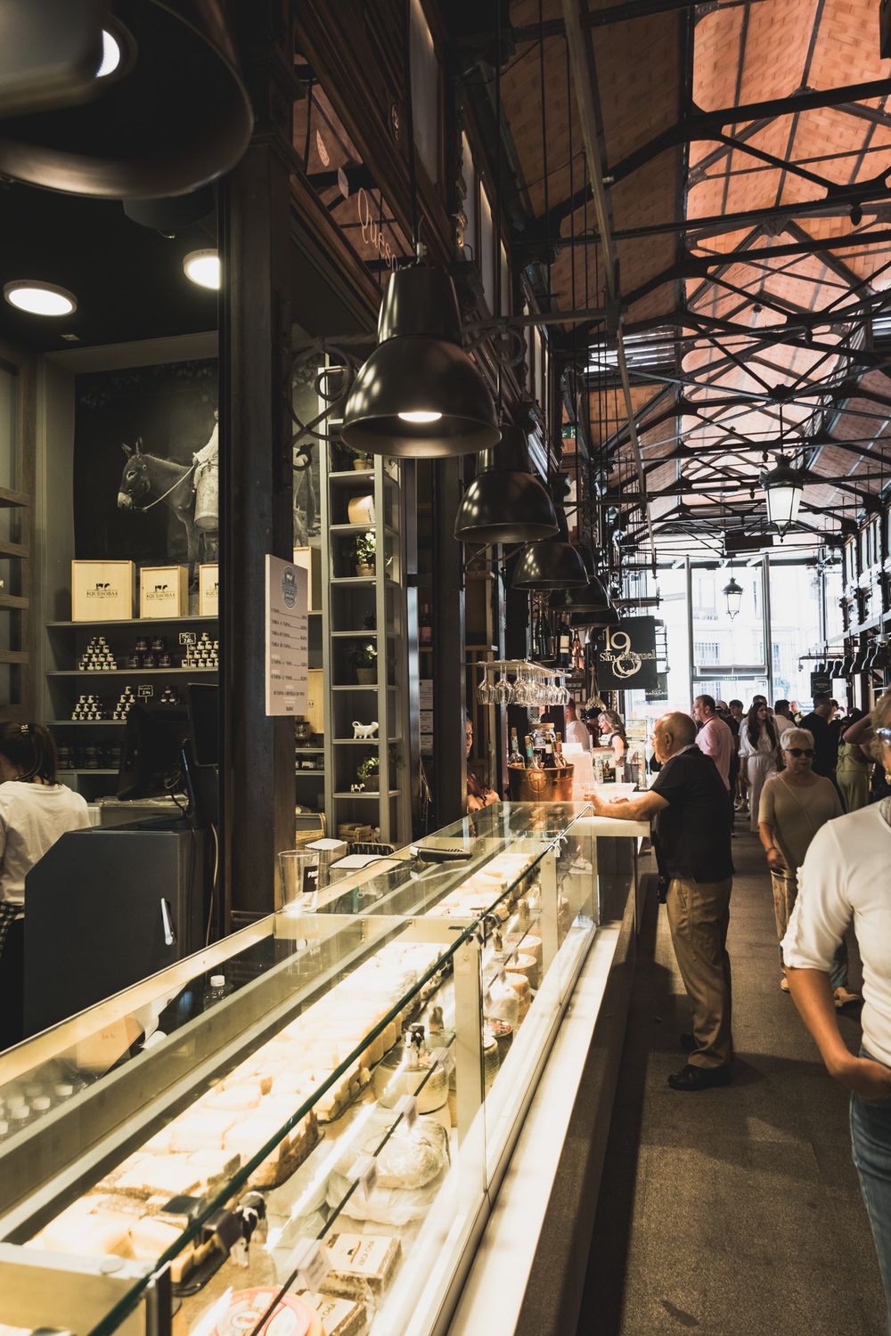 Crowded indoor gourmet market with long glass display counters, iron ceiling structure, and shoppers browsing food stalls.