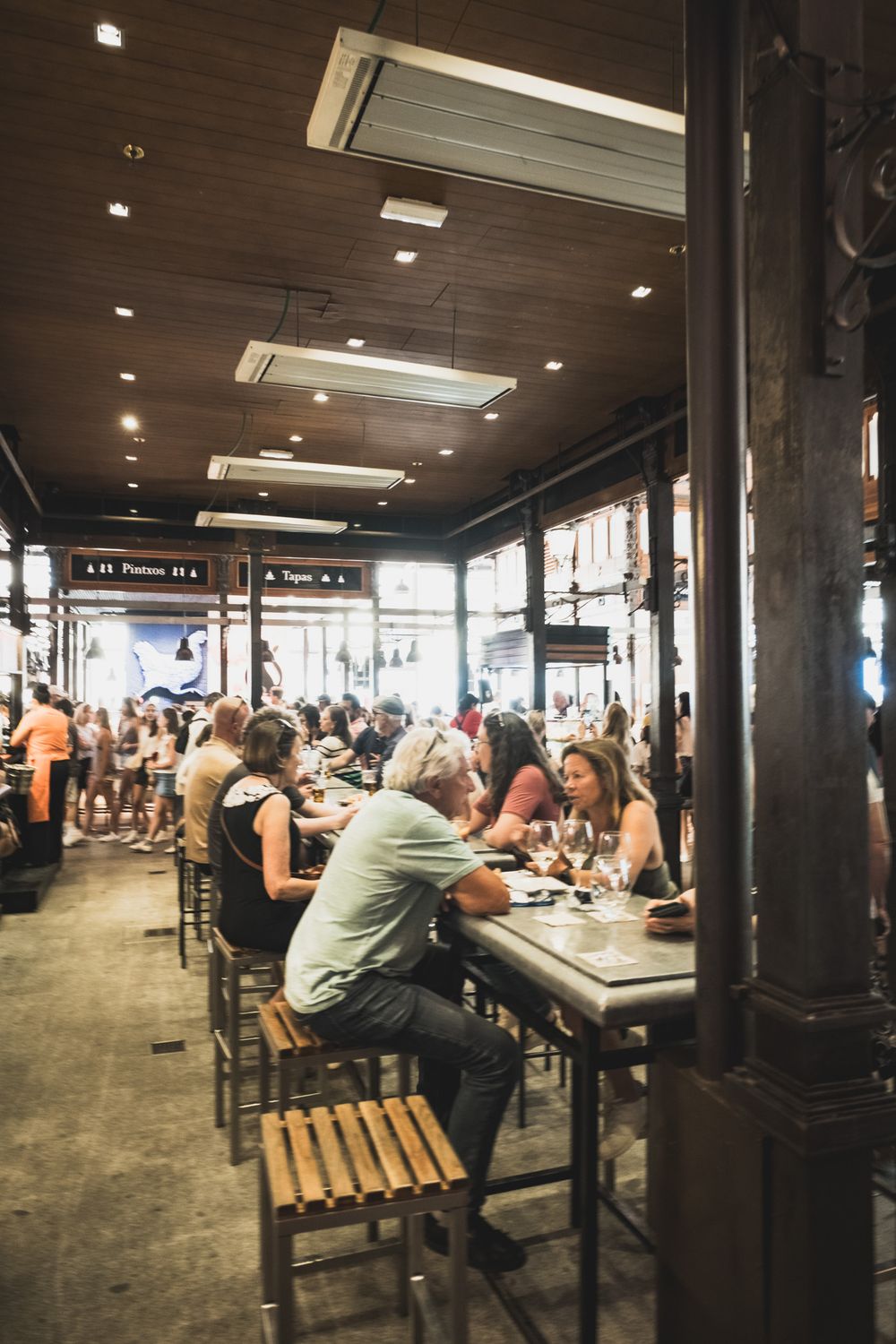 Diners seated at wooden bar tables enjoying food and drinks inside a lively historic market hall.
