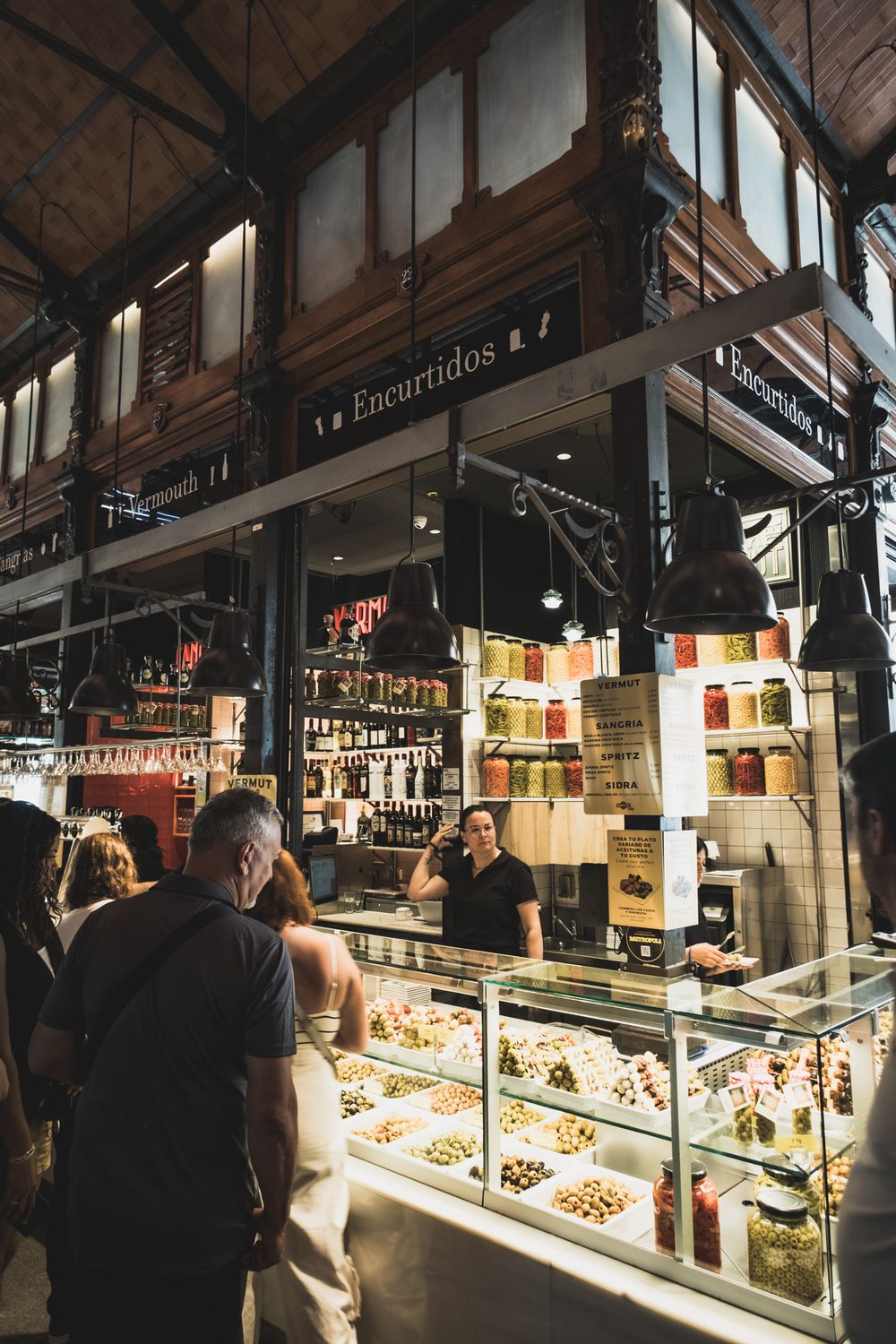 Encurtidos pickled goods stall with olives and preserved foods at a bustling Spanish indoor market.