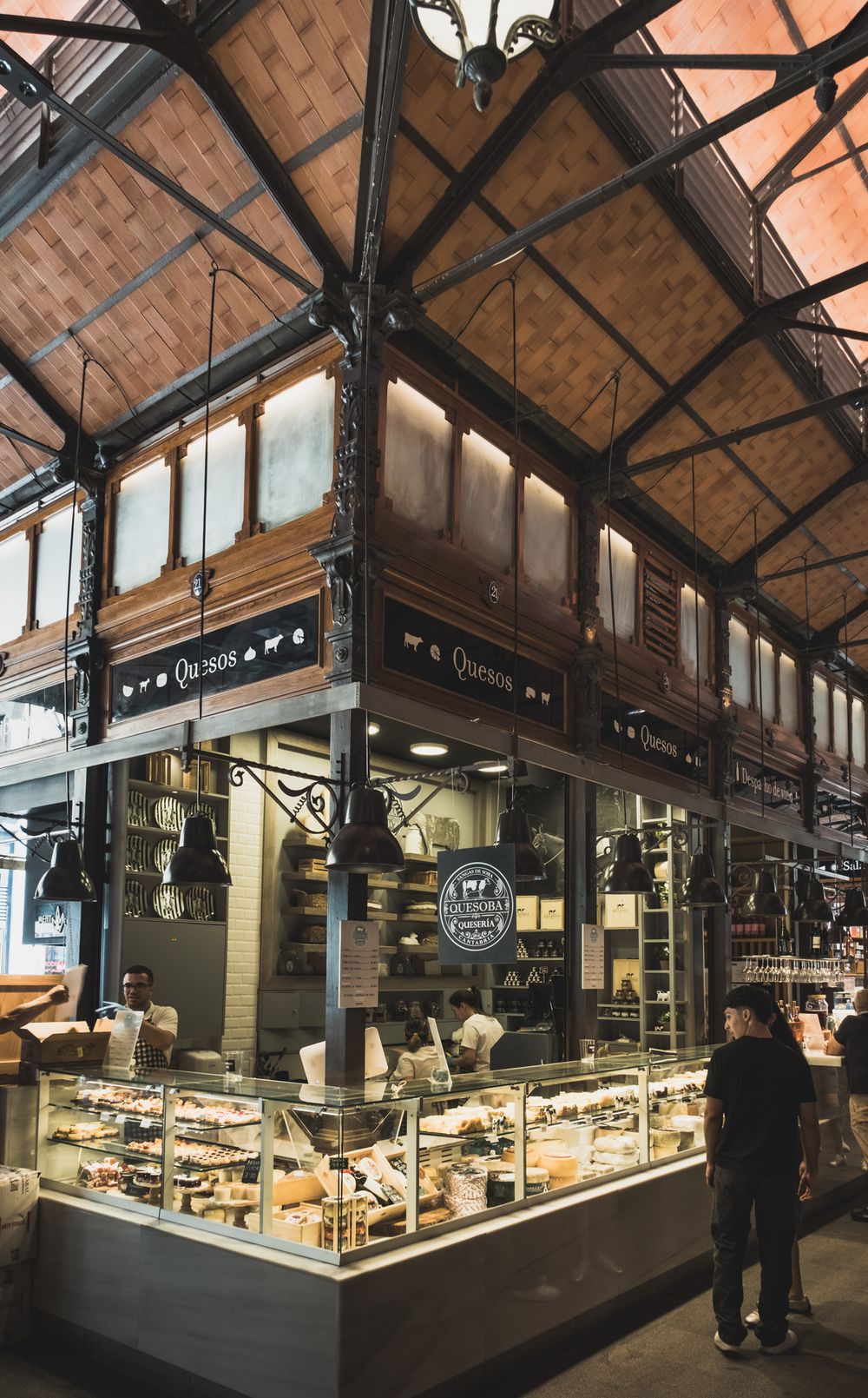 Quesos cheese shop stall with glass display cases inside a grand iron-roofed Spanish food market.
