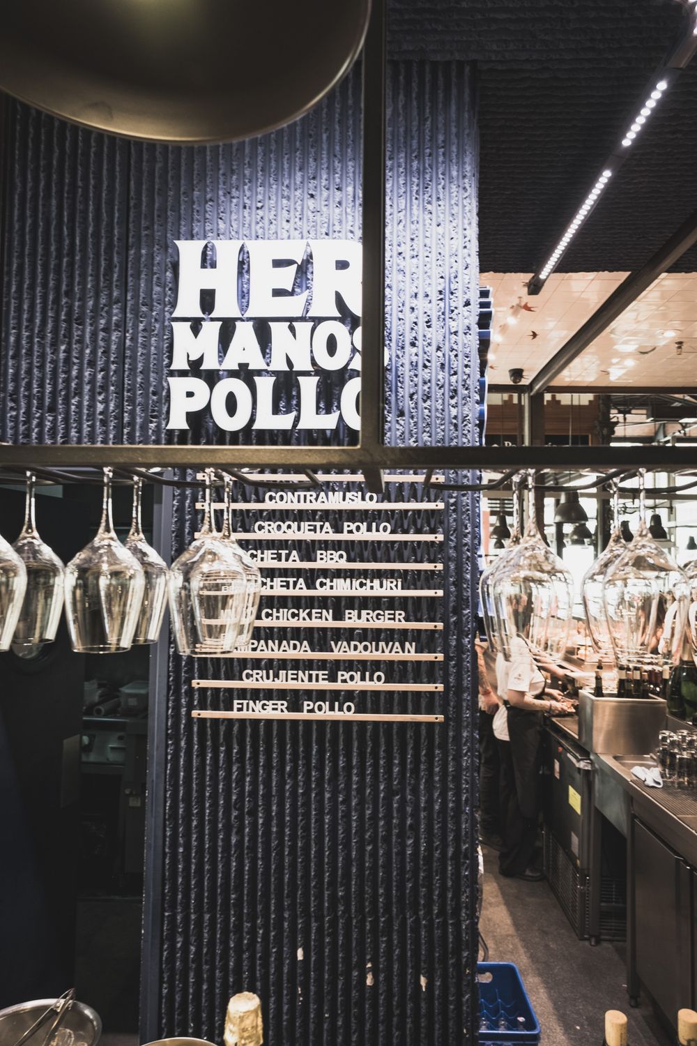 Hermanos Pollo restaurant menu board with hanging wine glasses displaying Spanish chicken dishes in a dark interior.