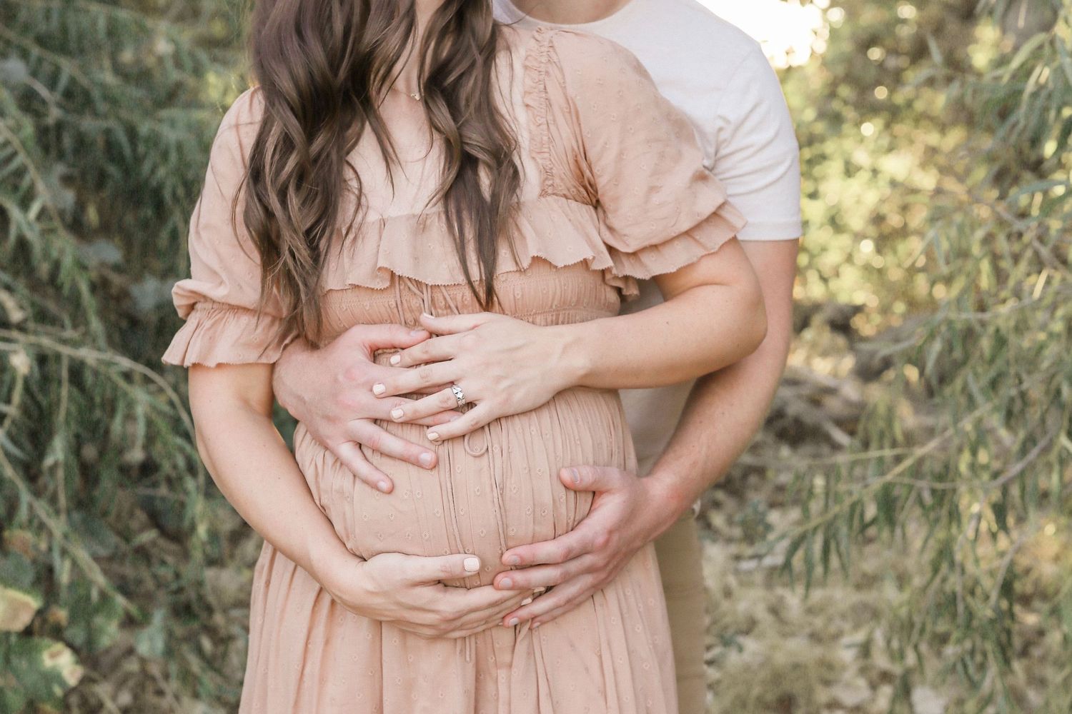 Pregnant woman in blush ruffle dress cradled by husband outdoors in a tender maternity photo session.