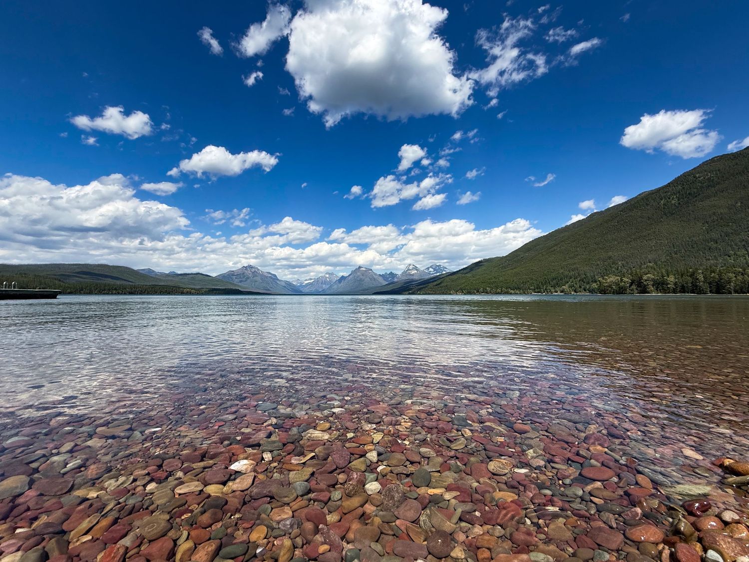 Crystal-clear shallow lake with colorful pebbles, mountain backdrop, and blue sky with clouds in Glacier National Park.