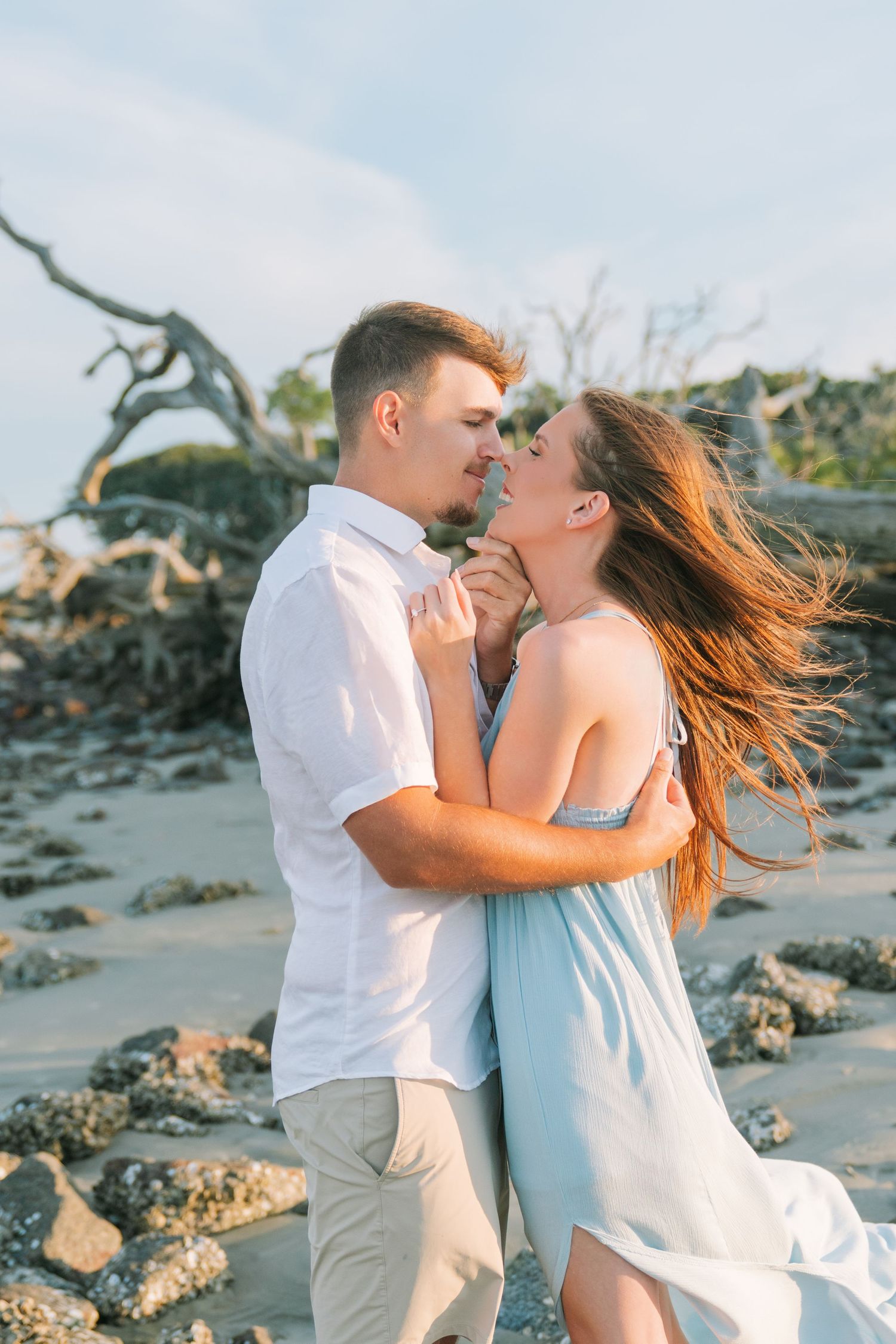 Couple embracing at Driftwood Beach on Jekyll Island with oak trees and rocks behind them after a surprise proposal engagement session