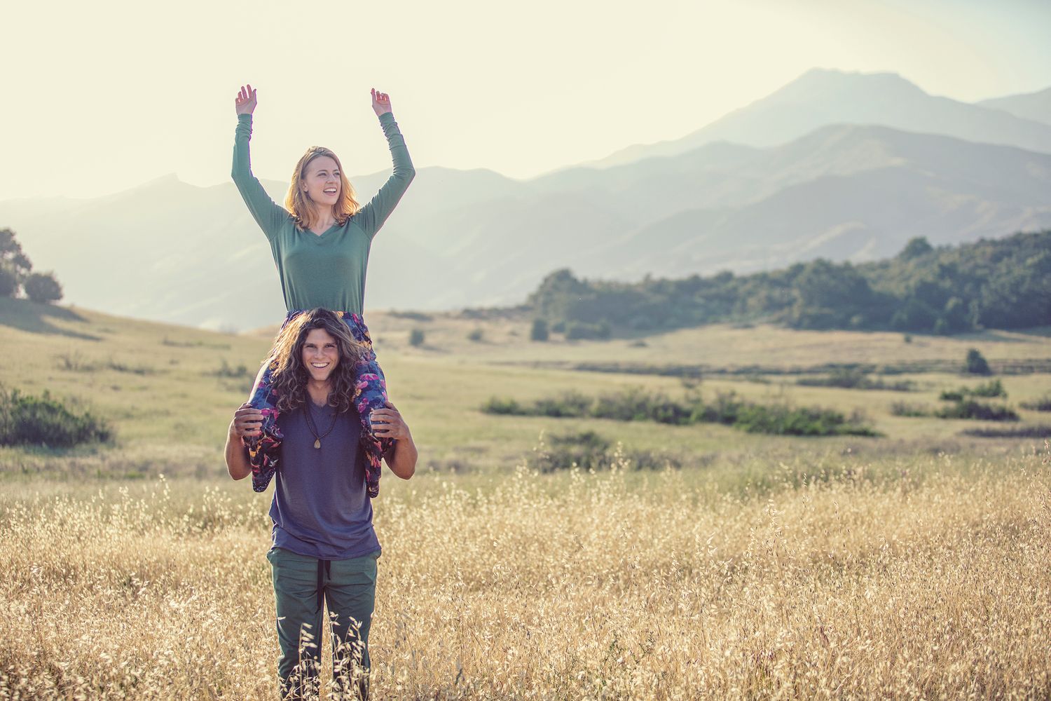 A woman sits on a man's shoulders with arms raised in a golden meadow with mountain range backdrop.
