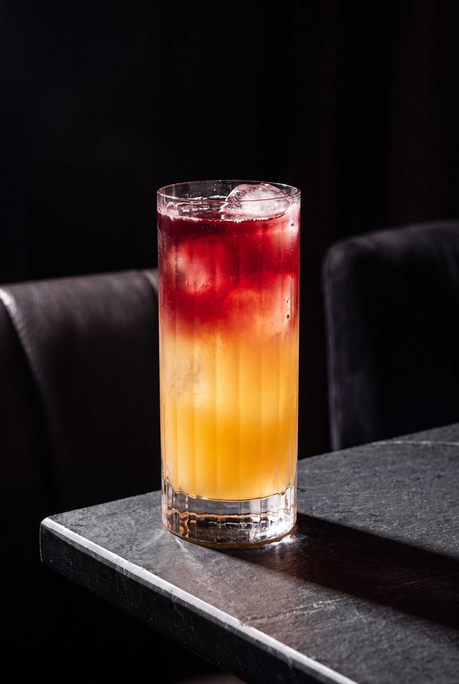 A layered cocktail with red and orange hues in a tall glass with ice, set on a dark stone surface.