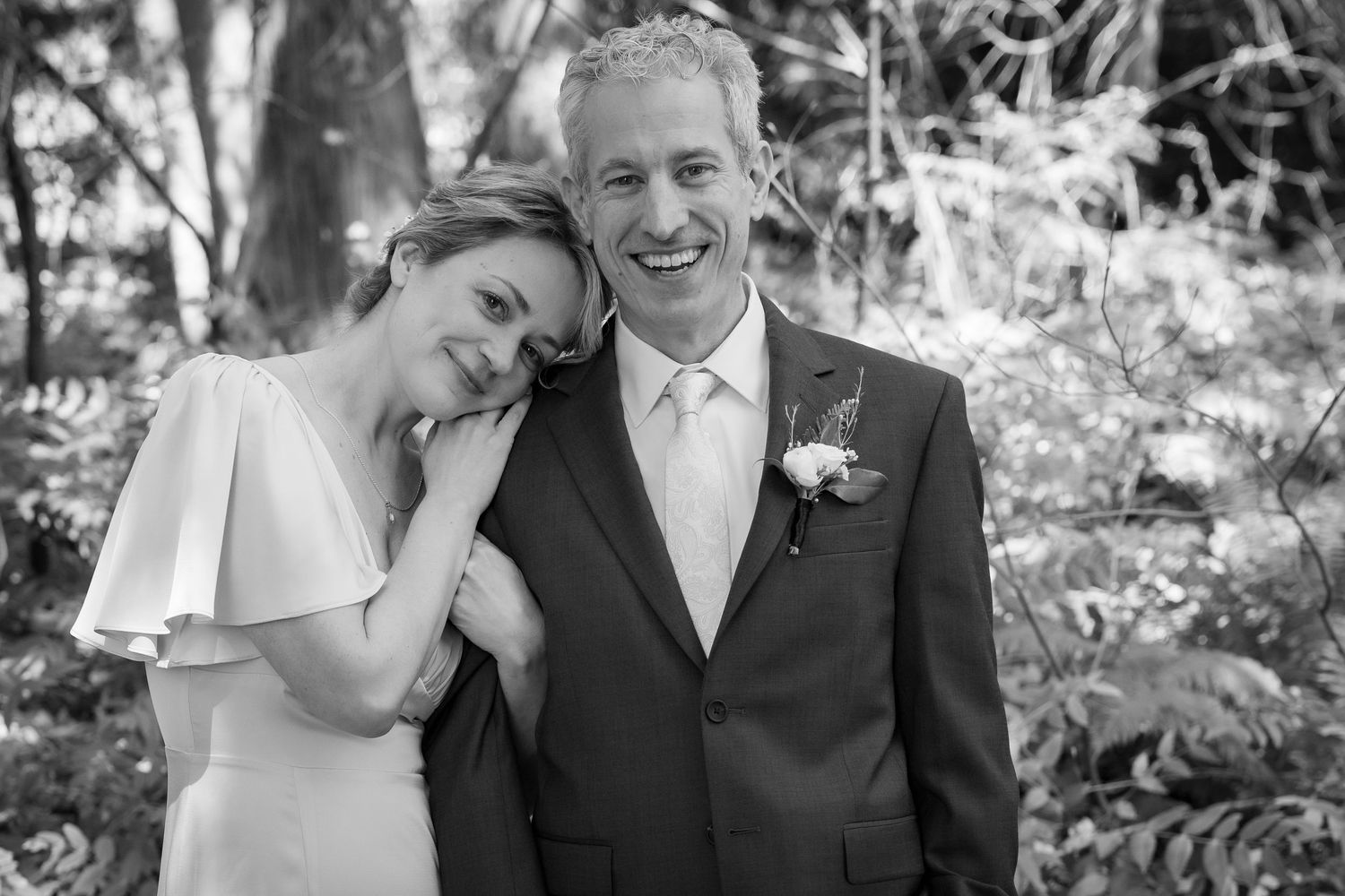 Black and white wedding photo of smiling bride and groom posing together outdoors among lush greenery.