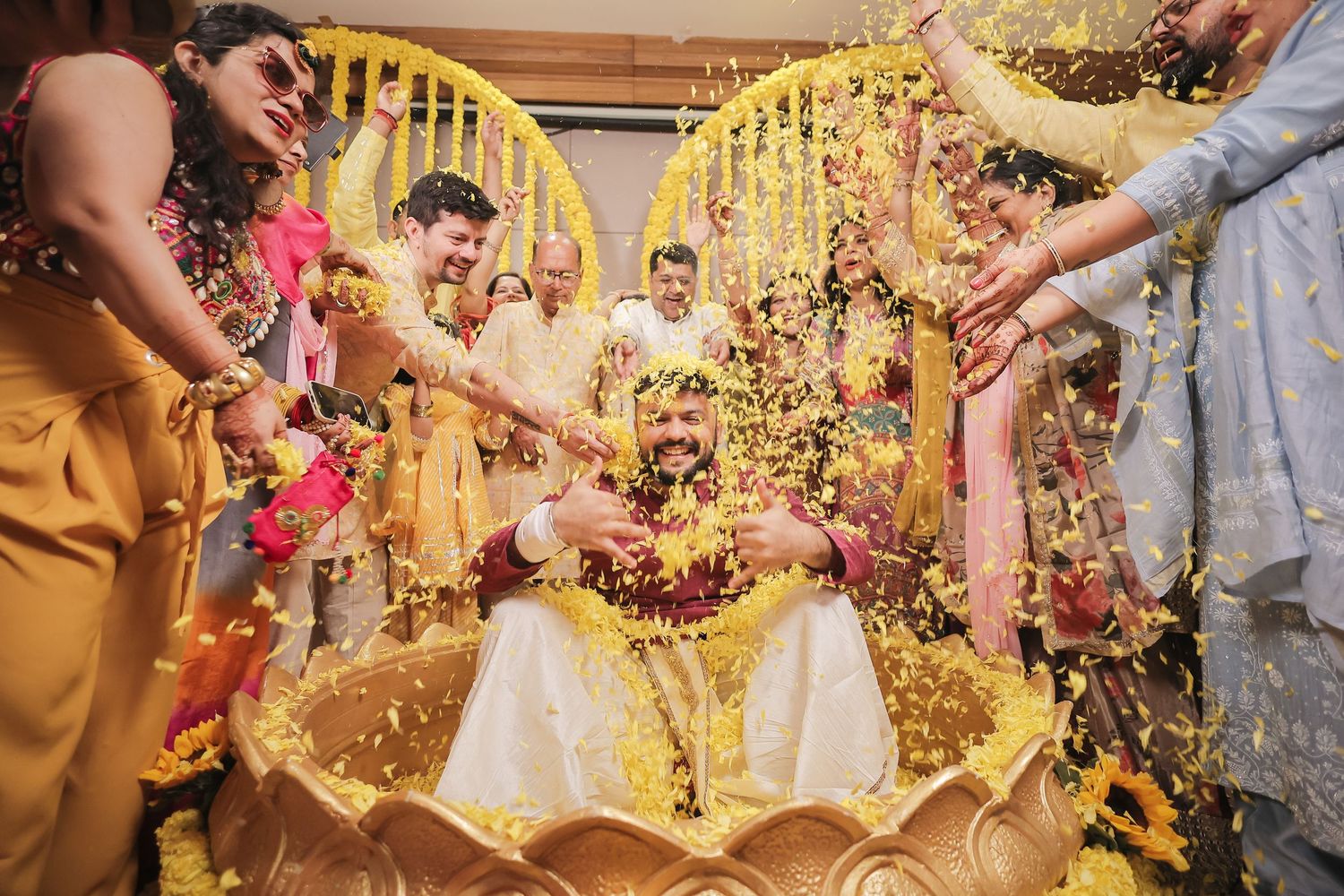 Colorful Indian wedding ceremony with couple seated in golden vessel, surrounded by guests showering flower petals.