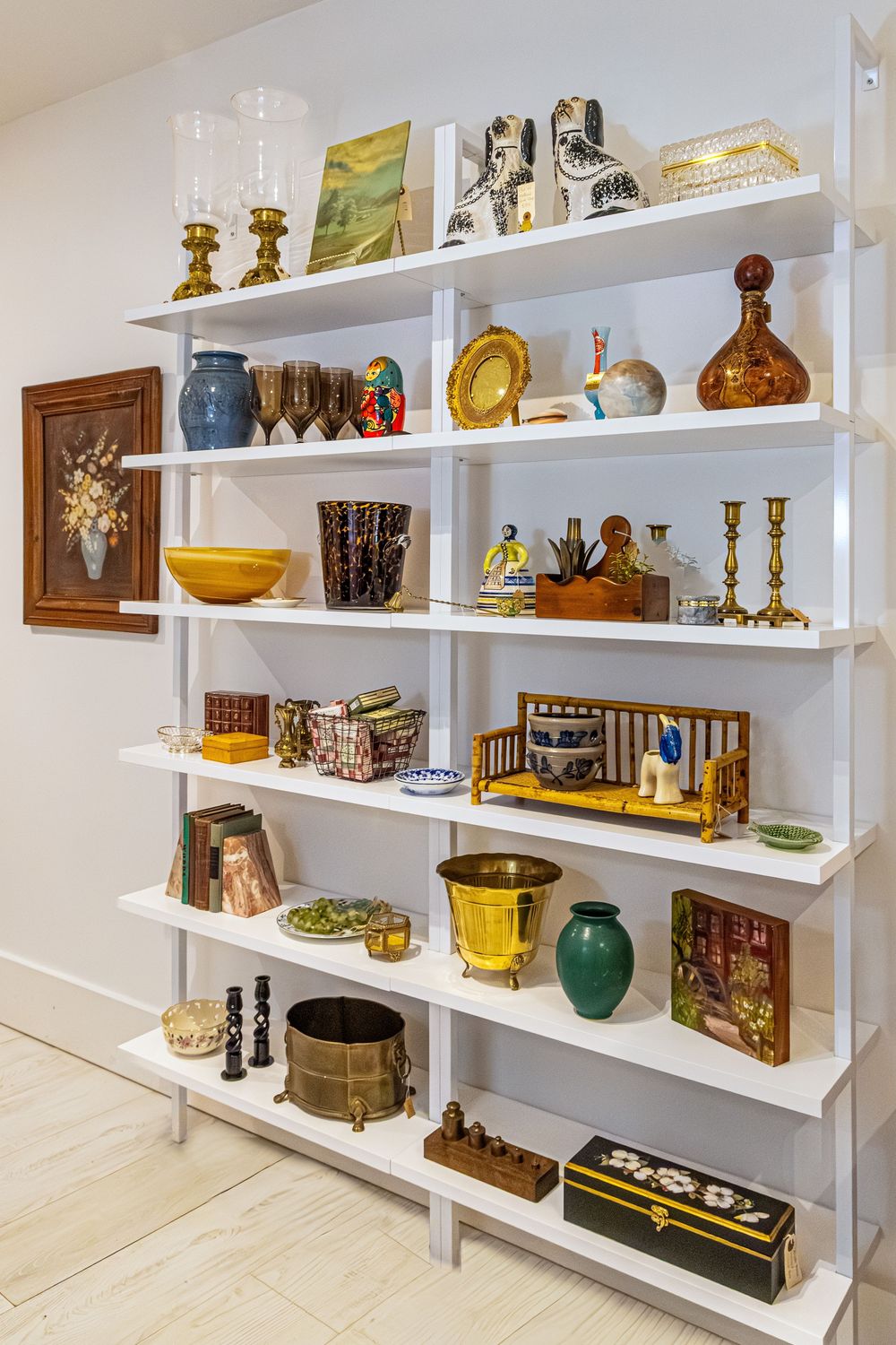 White shelving unit displaying colorful vintage decor including brass candlesticks, ceramic vases, glassware, and decorative boxes.