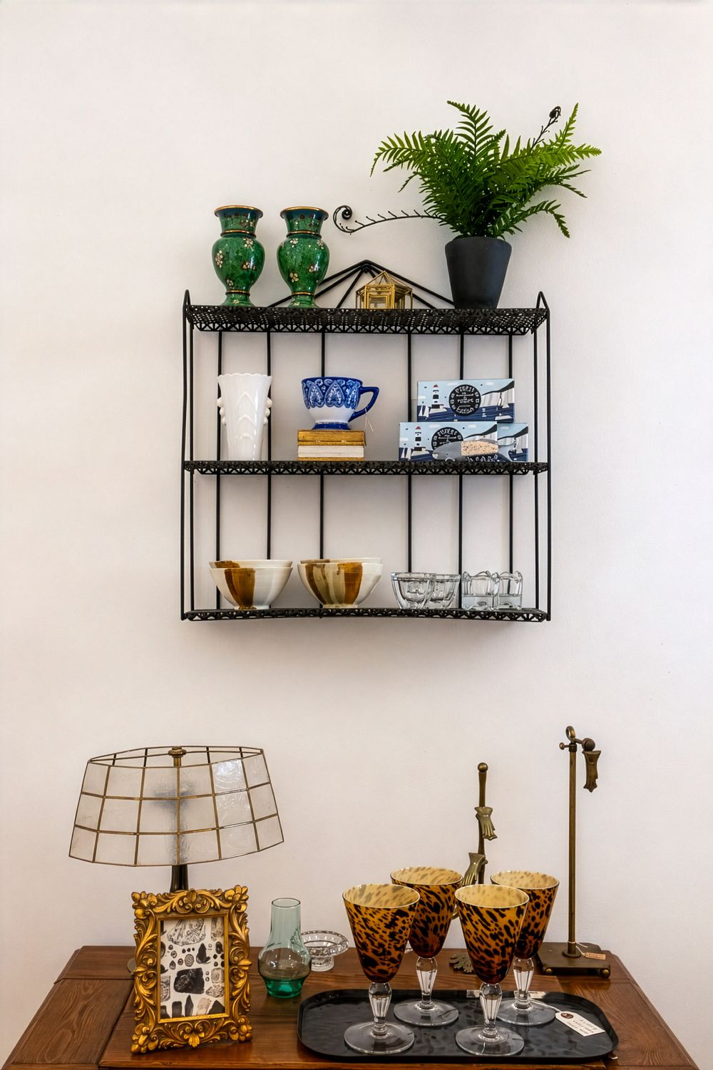 Vintage wall shelf with green vases, fern plant, ceramics, above a wooden table with lamp, framed art, and amber glasses. Swell House