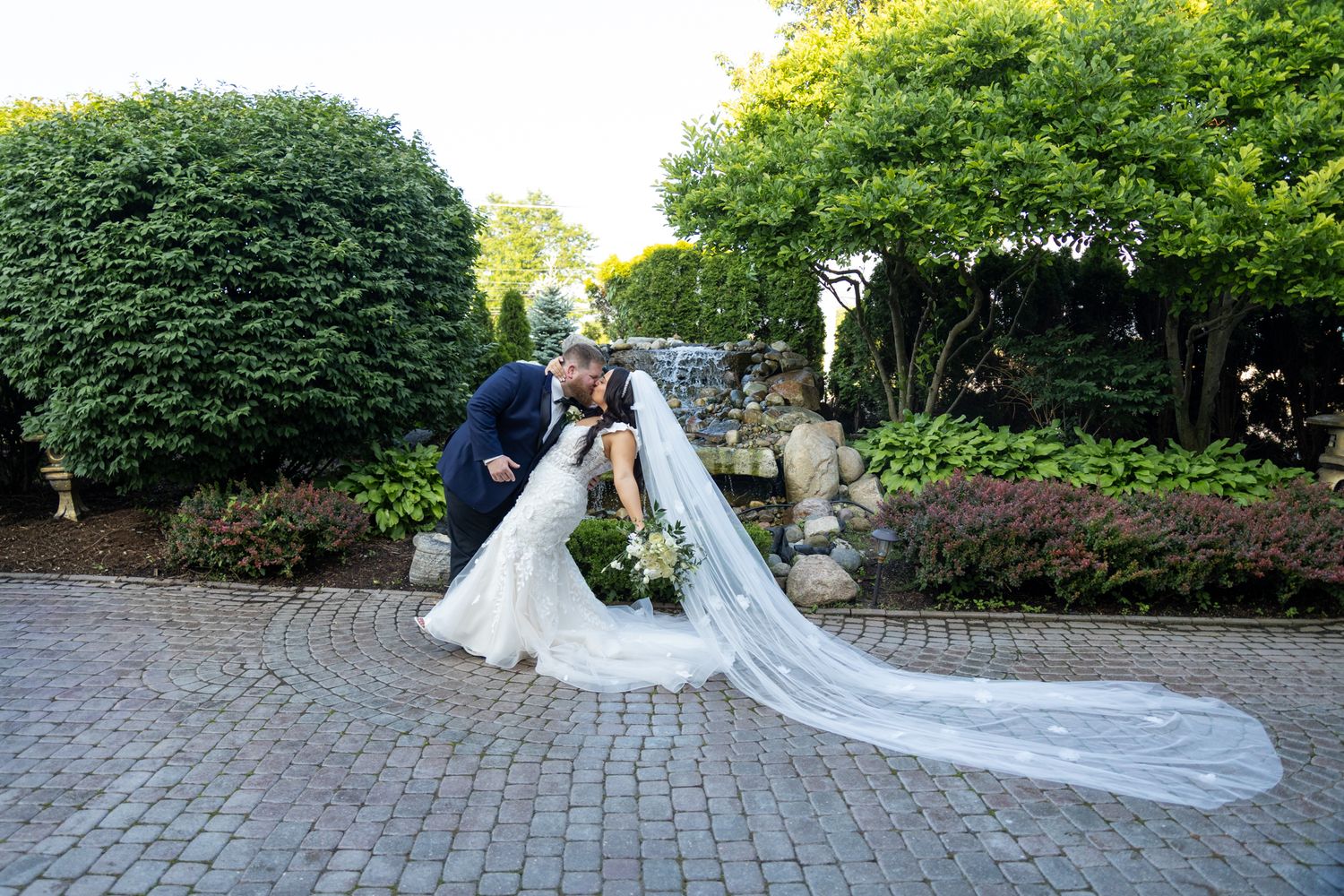 Bride and groom share a romantic dip kiss on a cobblestone path surrounded by lush greenery.