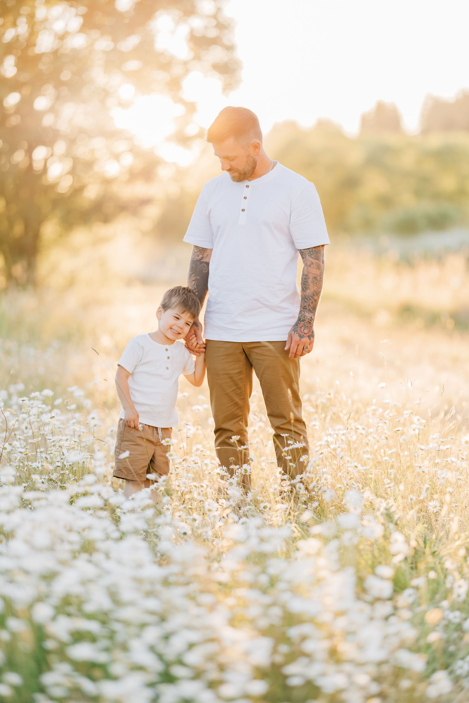 Father and young son holding hands in a sunlit daisy field, both wearing white shirts, sharing a tender moment.