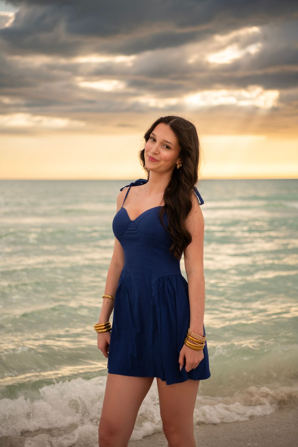 Smiling high school senior in blue dress posing on Clearwater Beach with dramatic sunset sky.