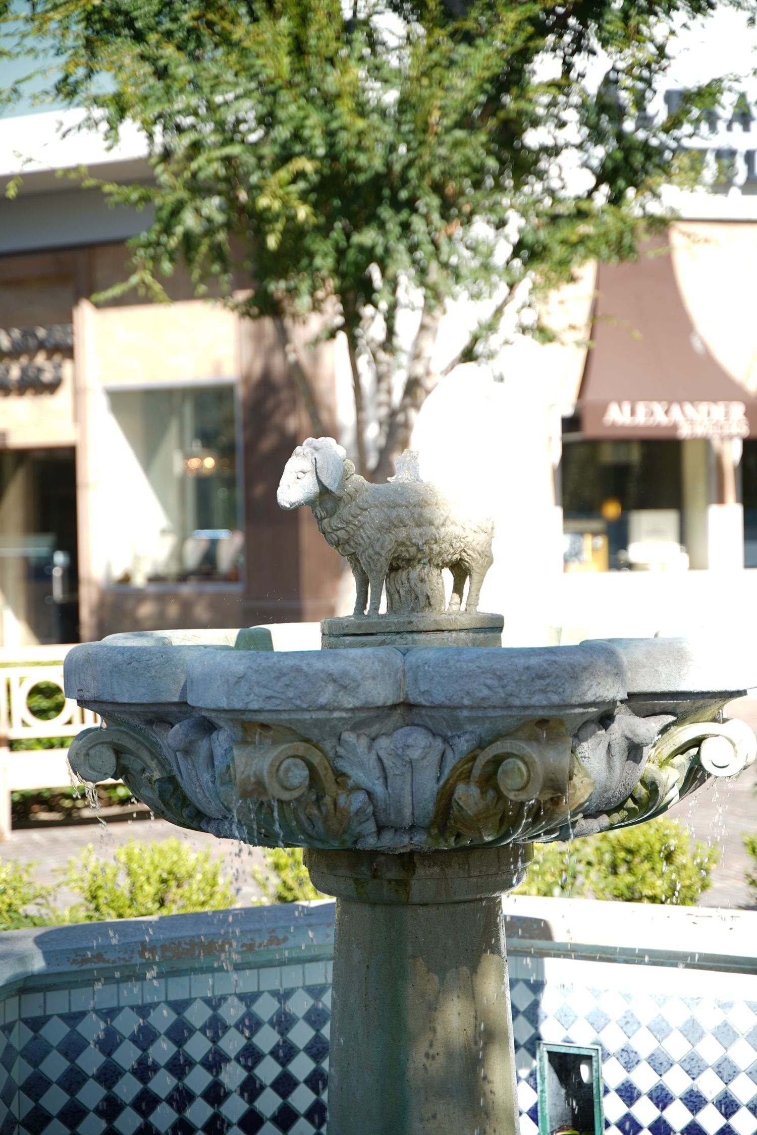 Ornate stone fountain topped with sheep sculpture in outdoor shopping plaza with trees and stores.