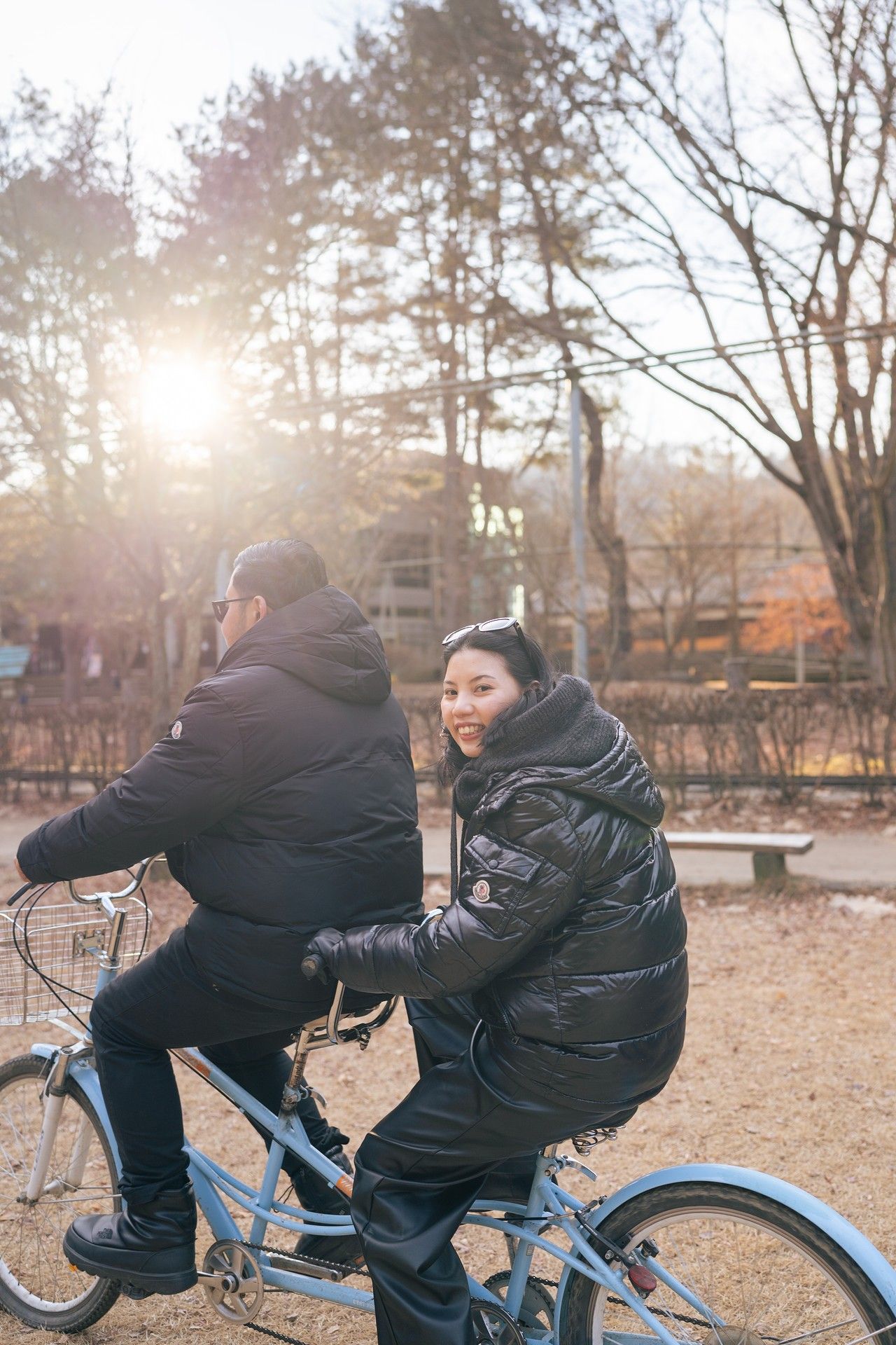 Couple riding a tandem bicycle in a park during golden hour, wearing black puffer jackets, with sunlight and bare trees behind them.