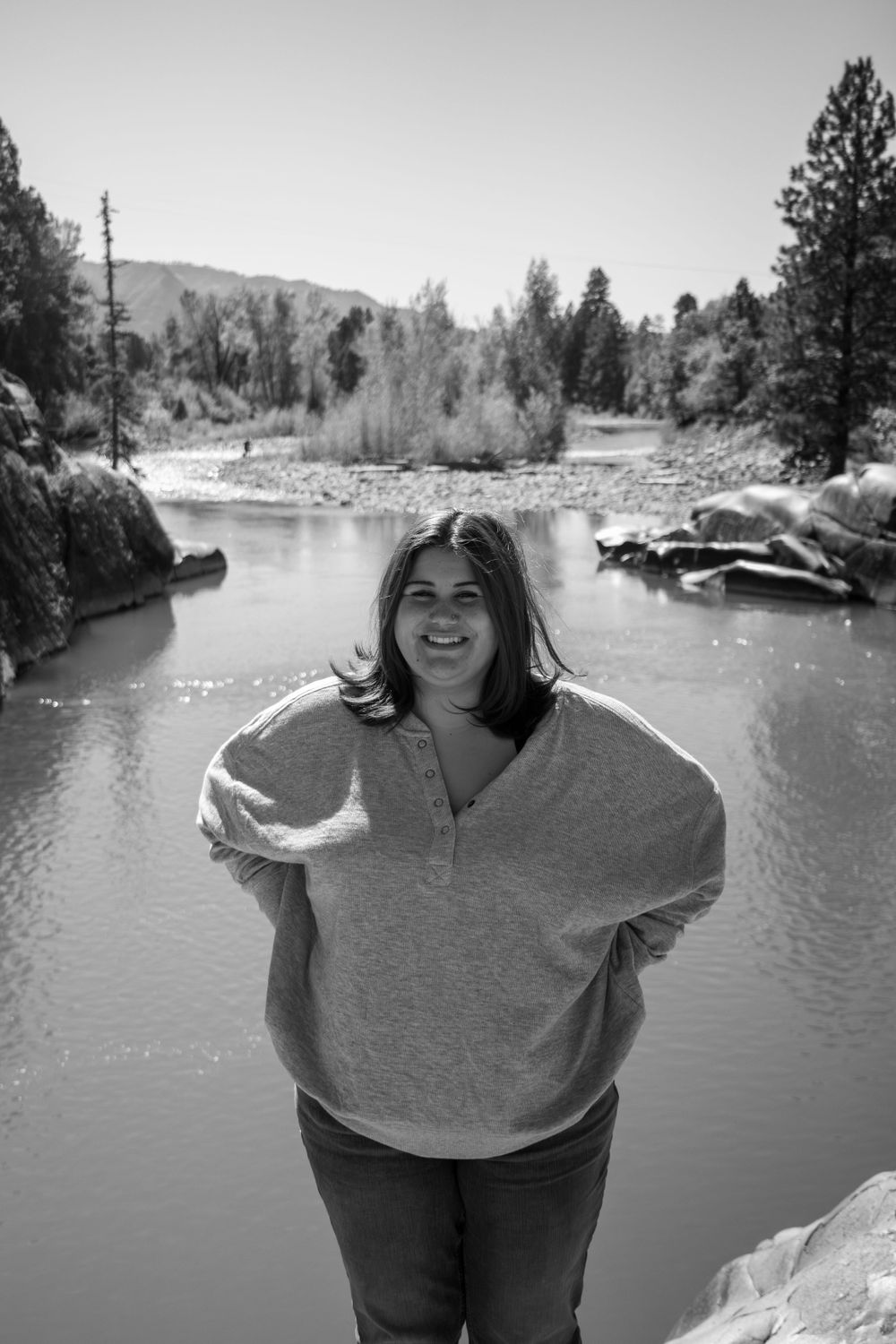 Black and white portrait by scenic river with mountains and trees in background.