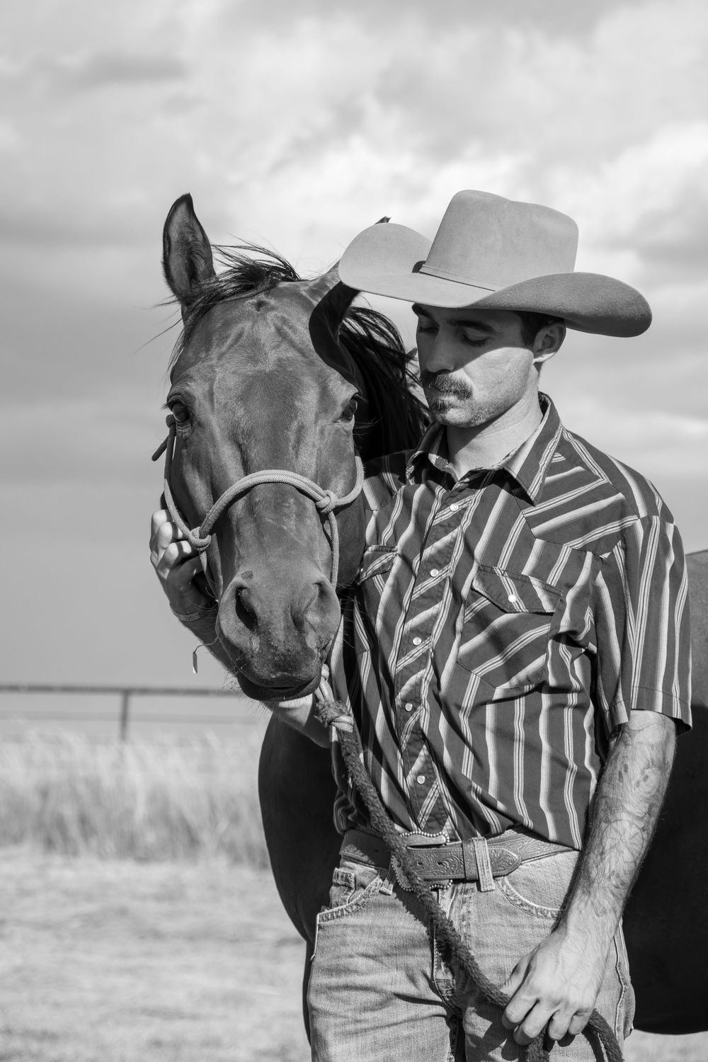 Black and white photo of a cowboy in a patterned shirt standing with a dark horse in an open field.