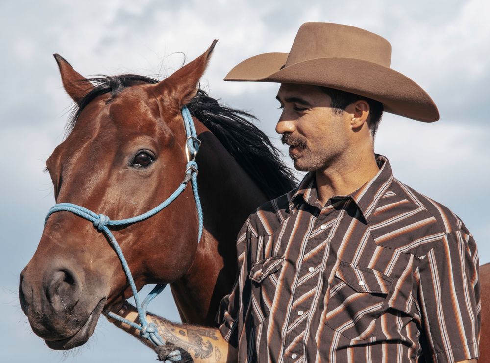 A cowboy in a brown striped shirt and wide-brimmed hat stands next to a chestnut horse with a blue halter.