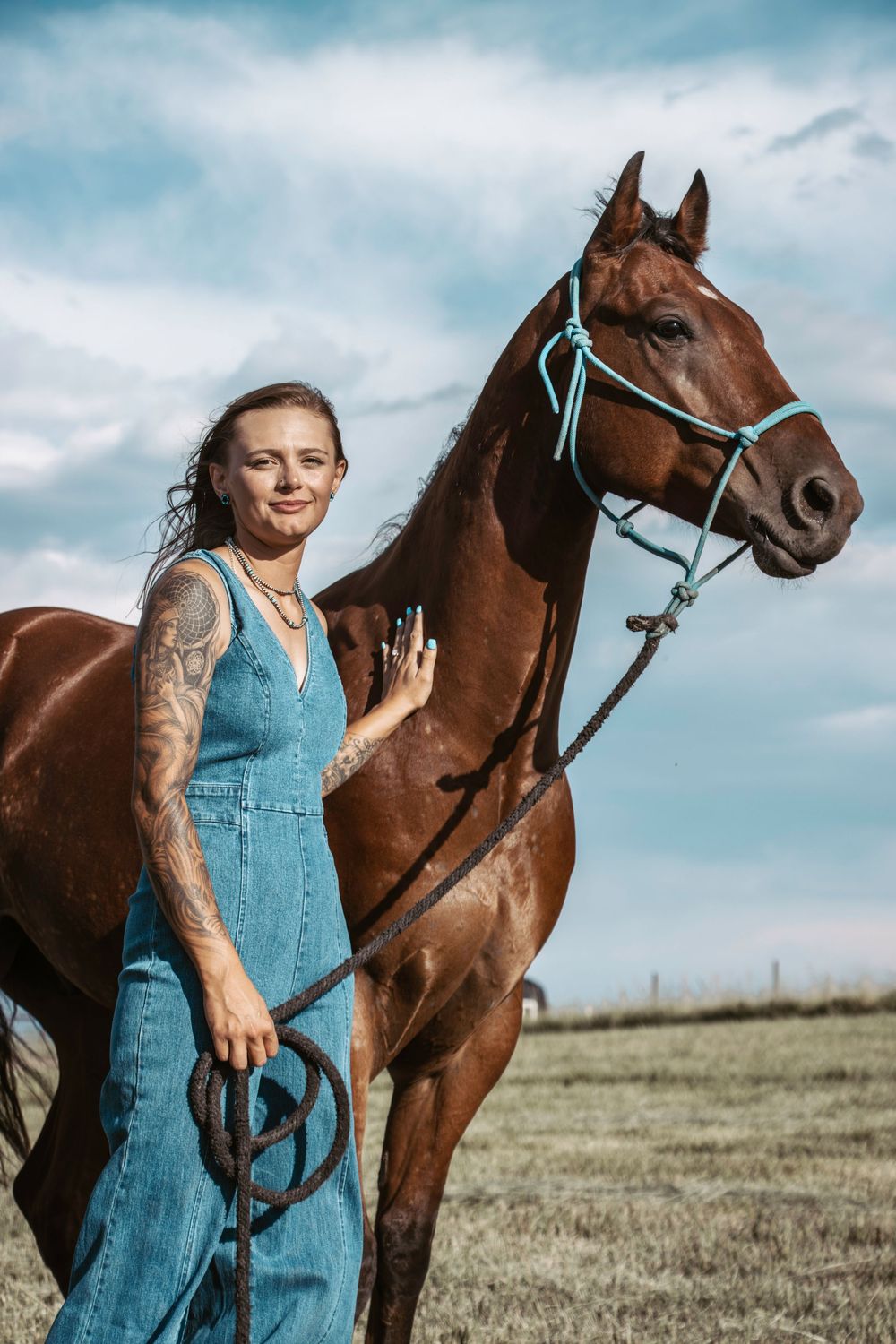 A brown horse stands in a grassy field next to someone wearing denim overalls on a cloudy day.