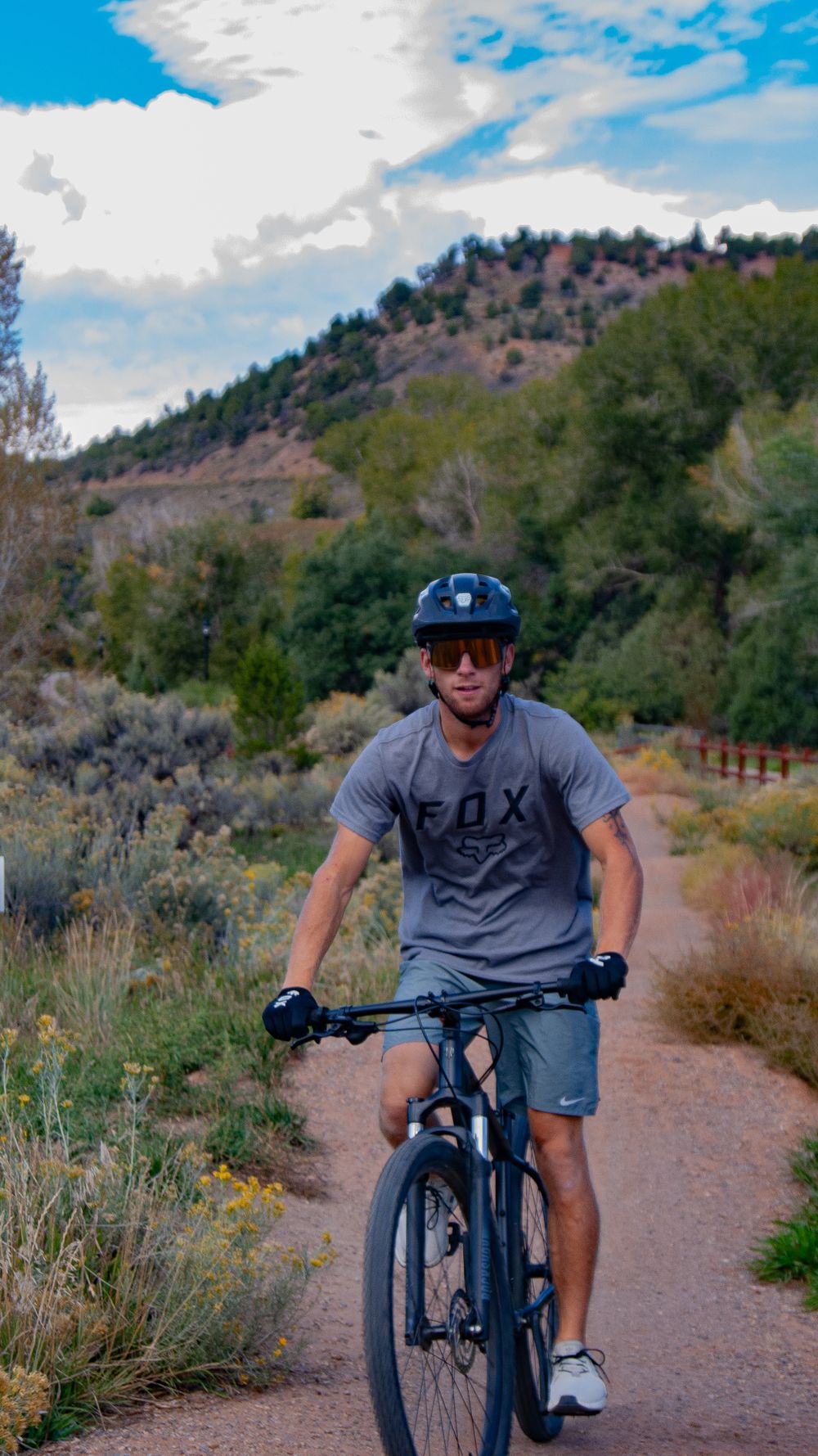 Cyclist poses on mountain bike along scenic desert trail with mountains and trees in background.