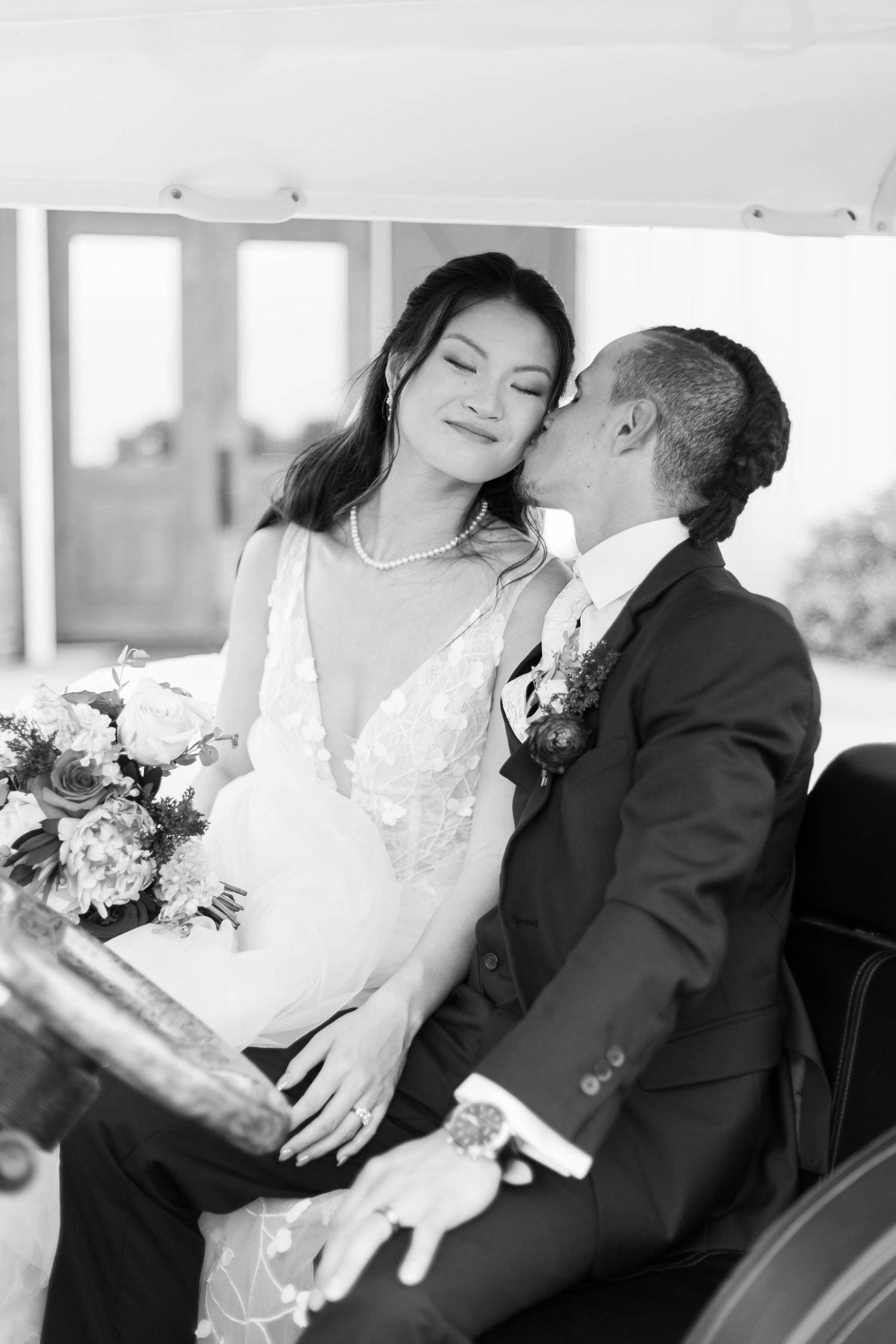 Black and white photo of a groom kissing bride on cheek, bride holding bouquet, seated in vintage car.