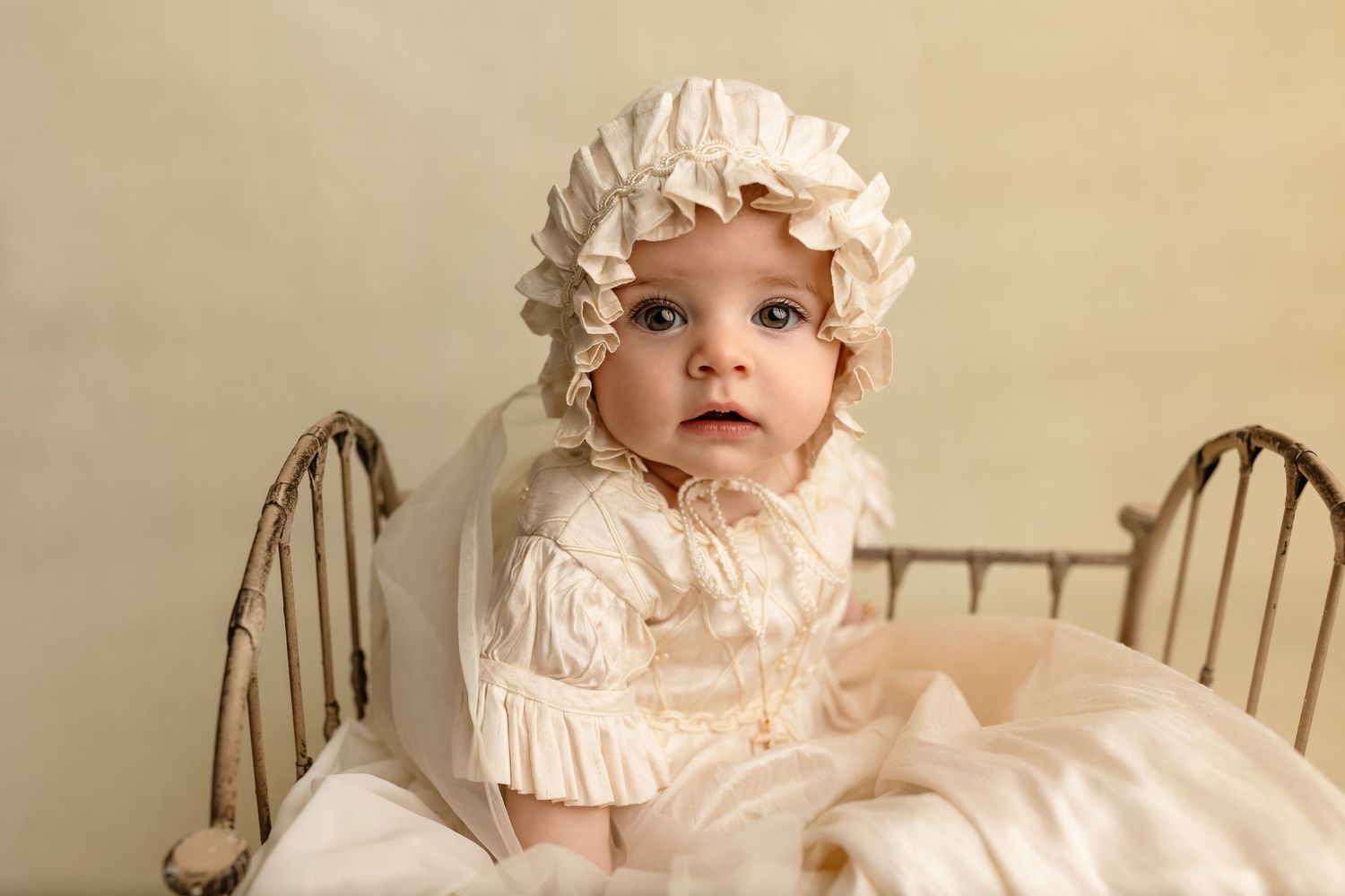 Gorgeous baby girl in a silk gown and bonnet sits in an antique crib for her milestone session by Charlottesville newborn photographer.
