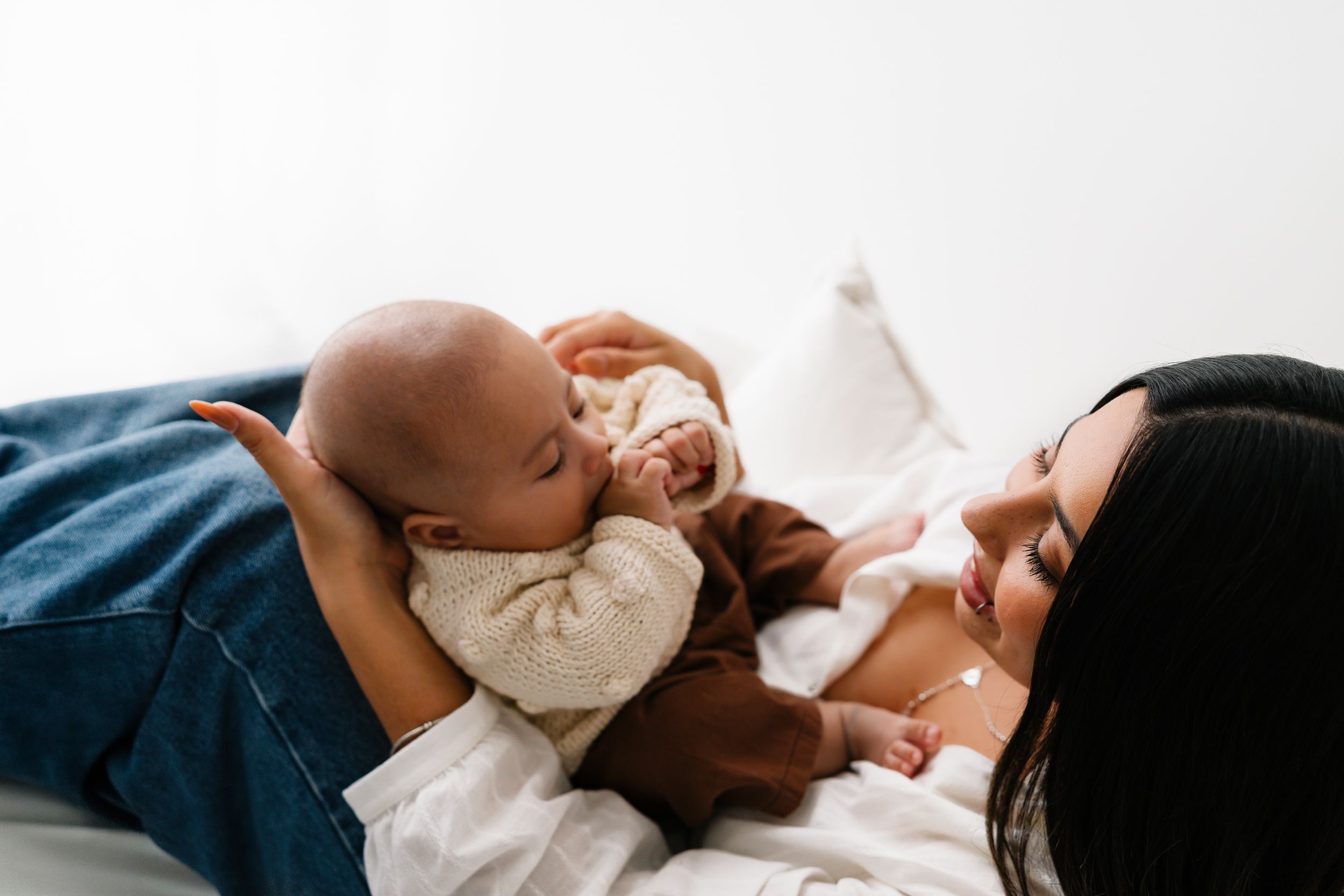 A mother lying down lovingly gazes at her newborn baby held by an older child on a white bed.