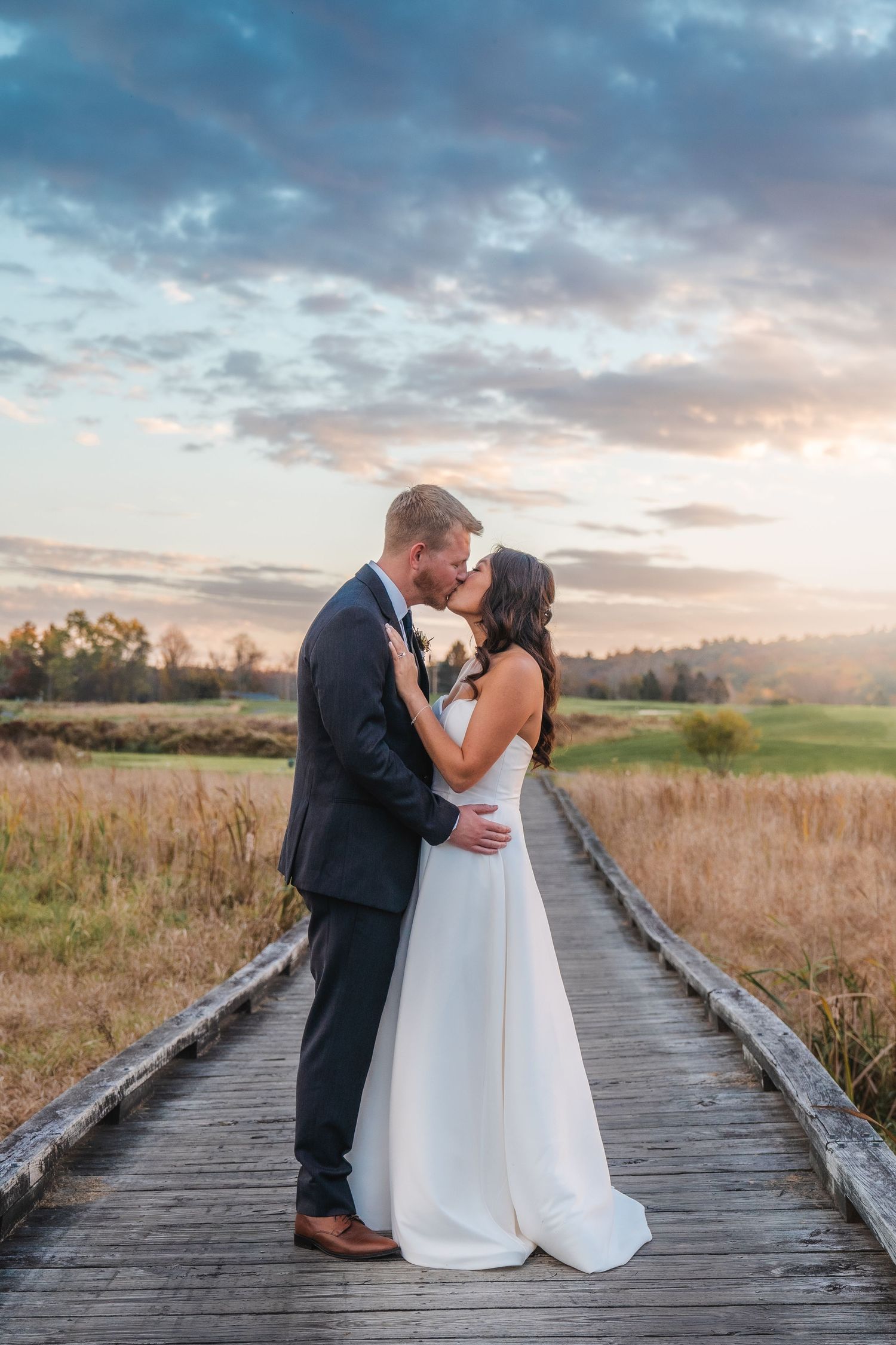 Newlywed couple sharing an intimate moment at sunset on a wooden boardwalk through a golden field.