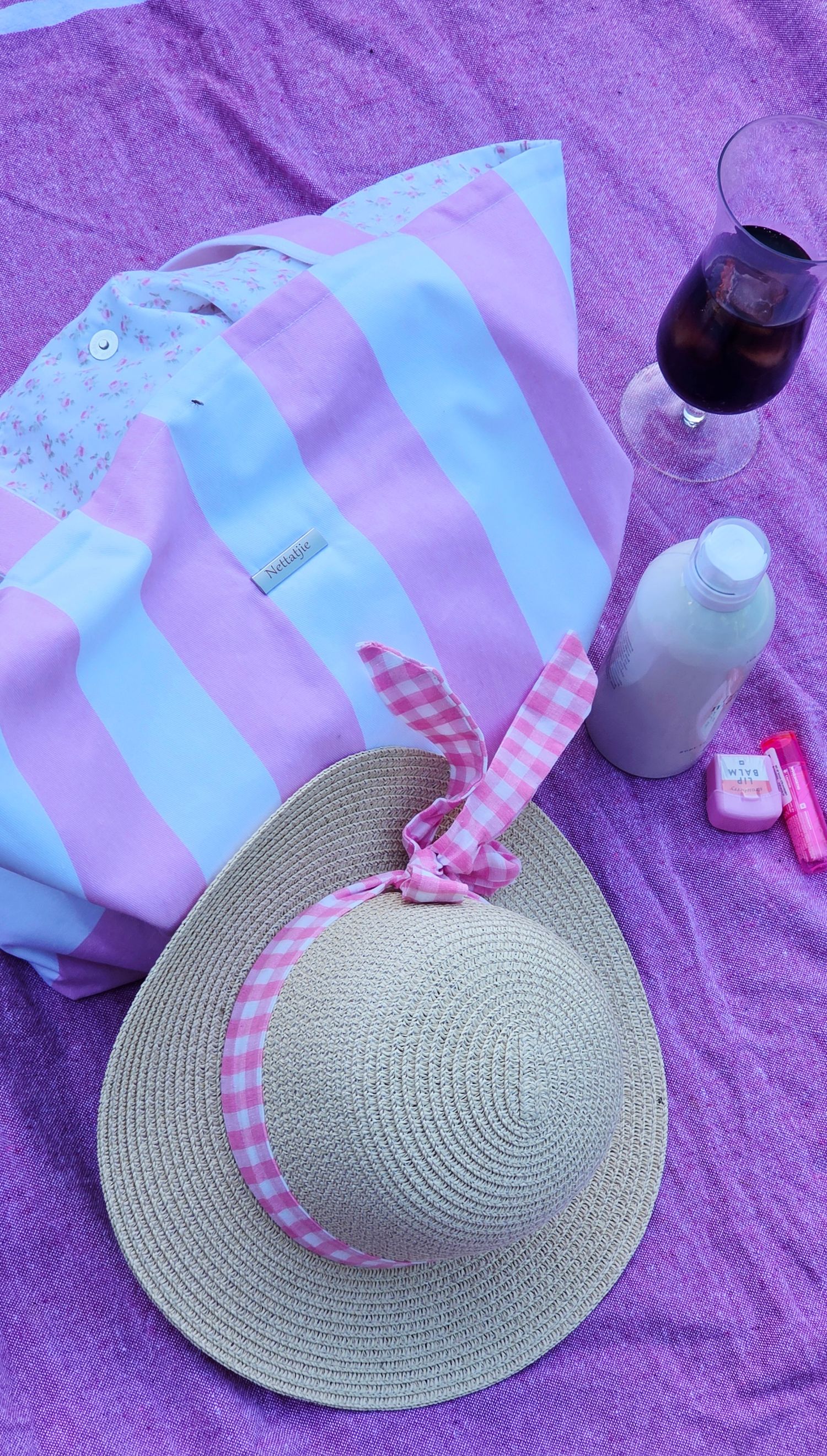 Pink striped beach tote bag, straw sun hat with gingham ribbon, wine glass, and sunscreen on purple blanket.