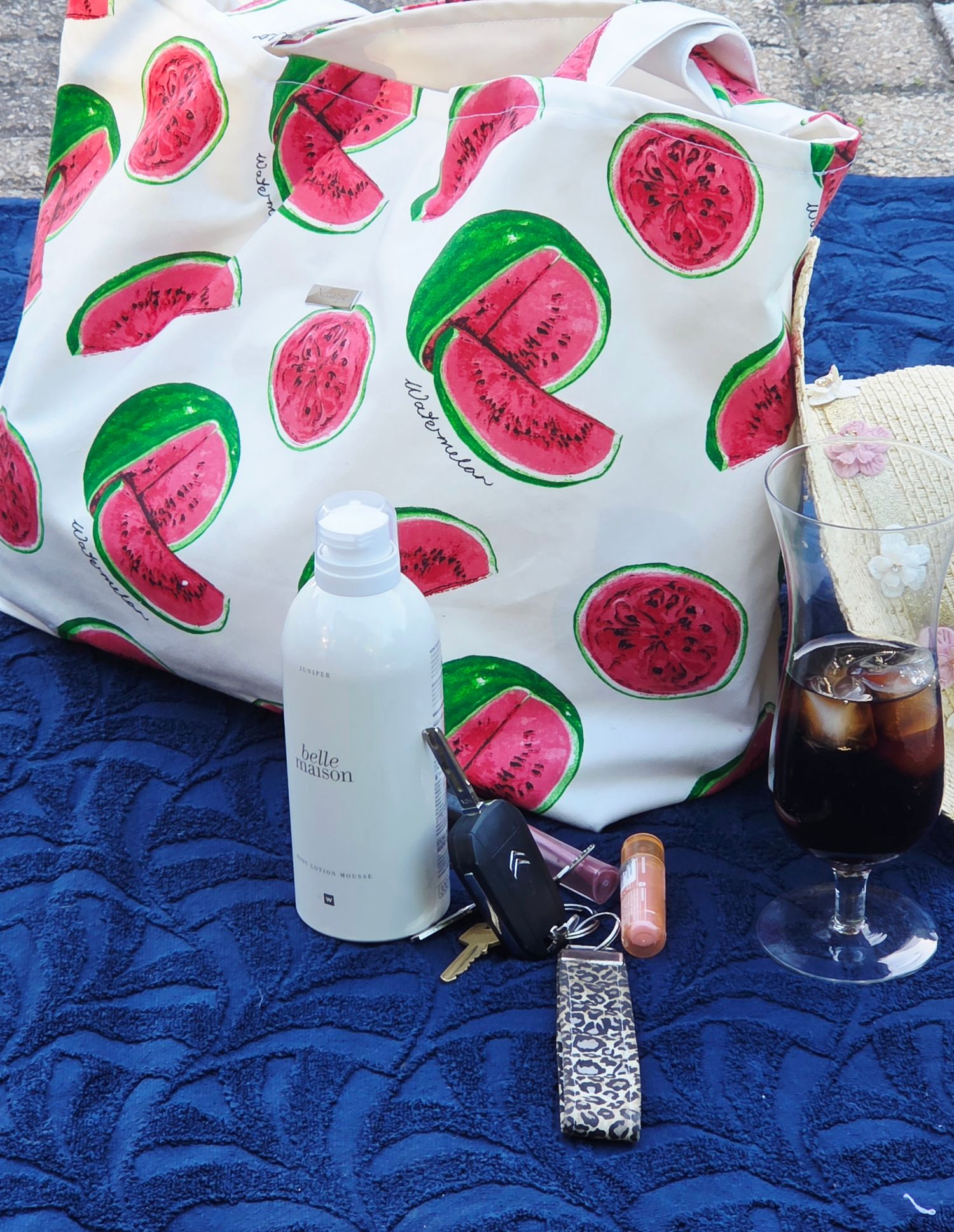 A watermelon-print tote bag on a blue towel with sunscreen, car keys, sunglasses, and a glass of iced cola.