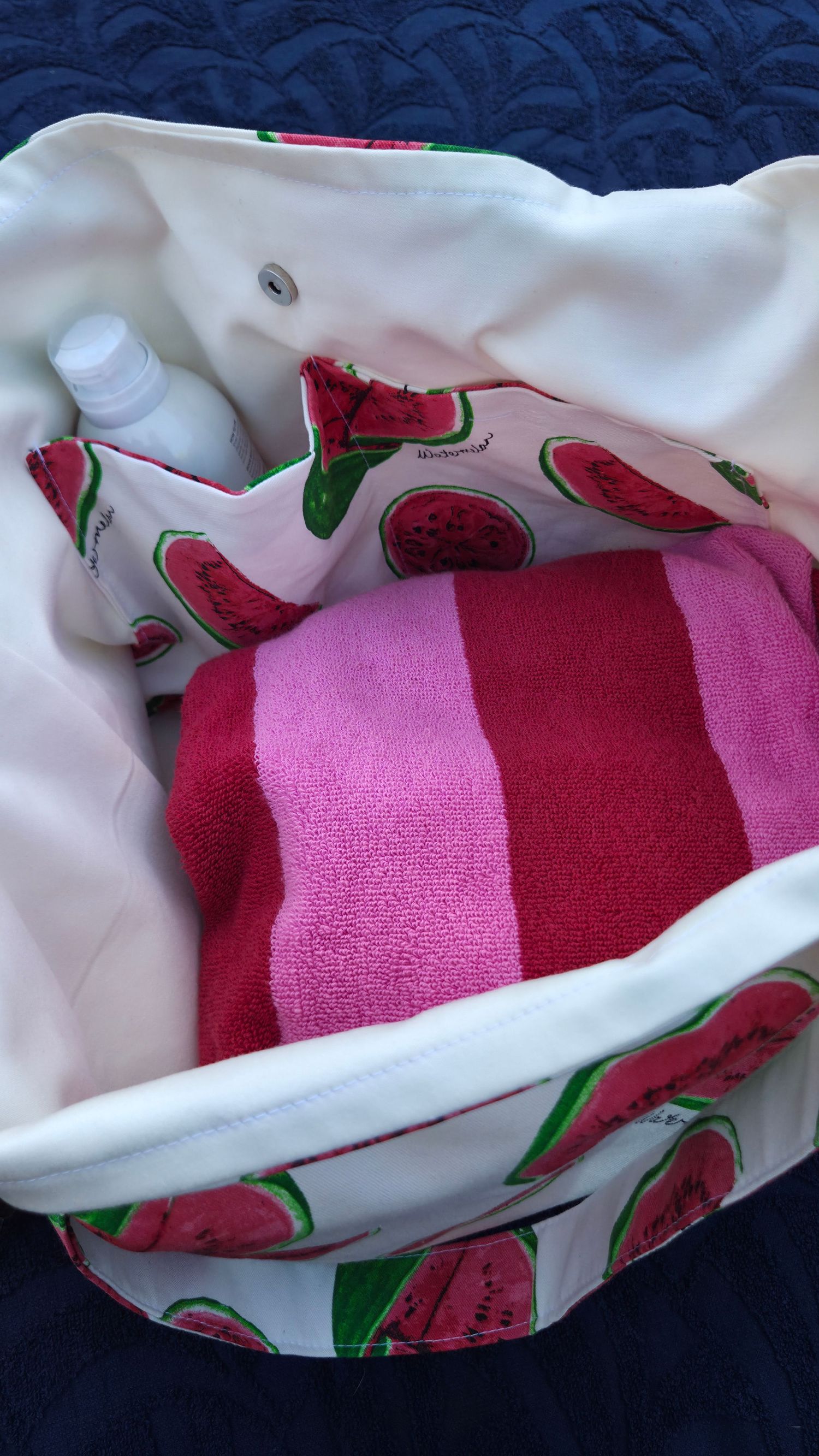 Watermelon print tote bag packed with a striped pink beach towel and sunscreen bottle.
