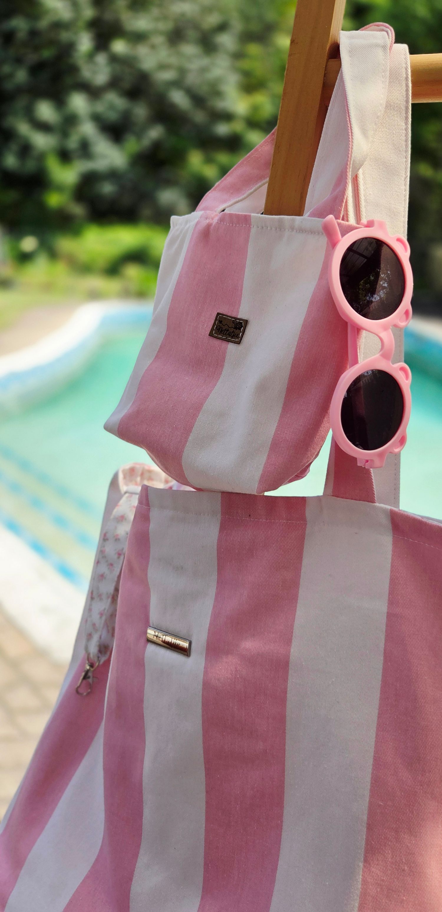 Pink and white striped beach tote bag with matching pouch and pink flower sunglasses hanging on a wooden rack near a pool.