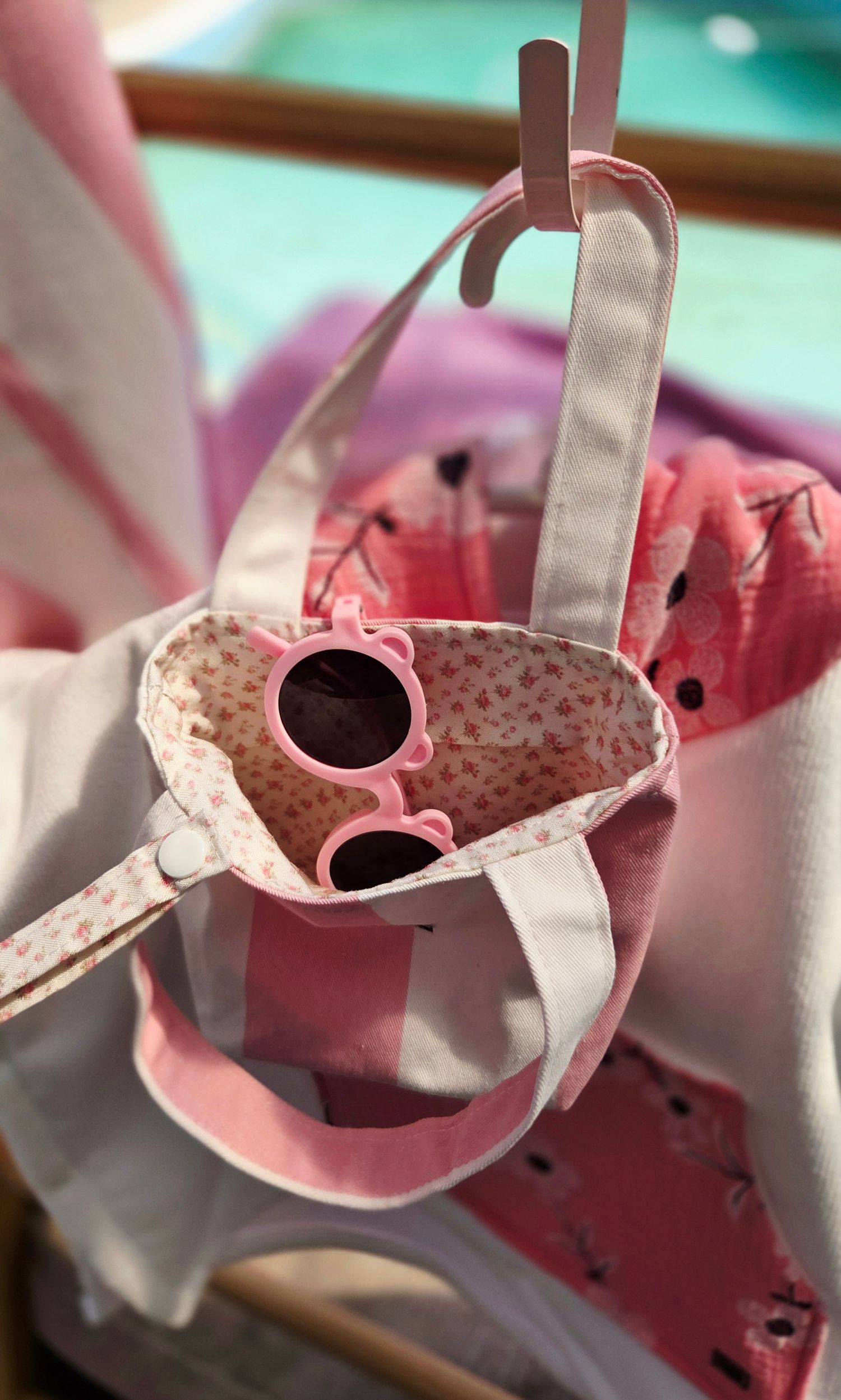 Pink floral tote bag with pink sunglasses inside, hanging on a rack with pink clothing in the background.