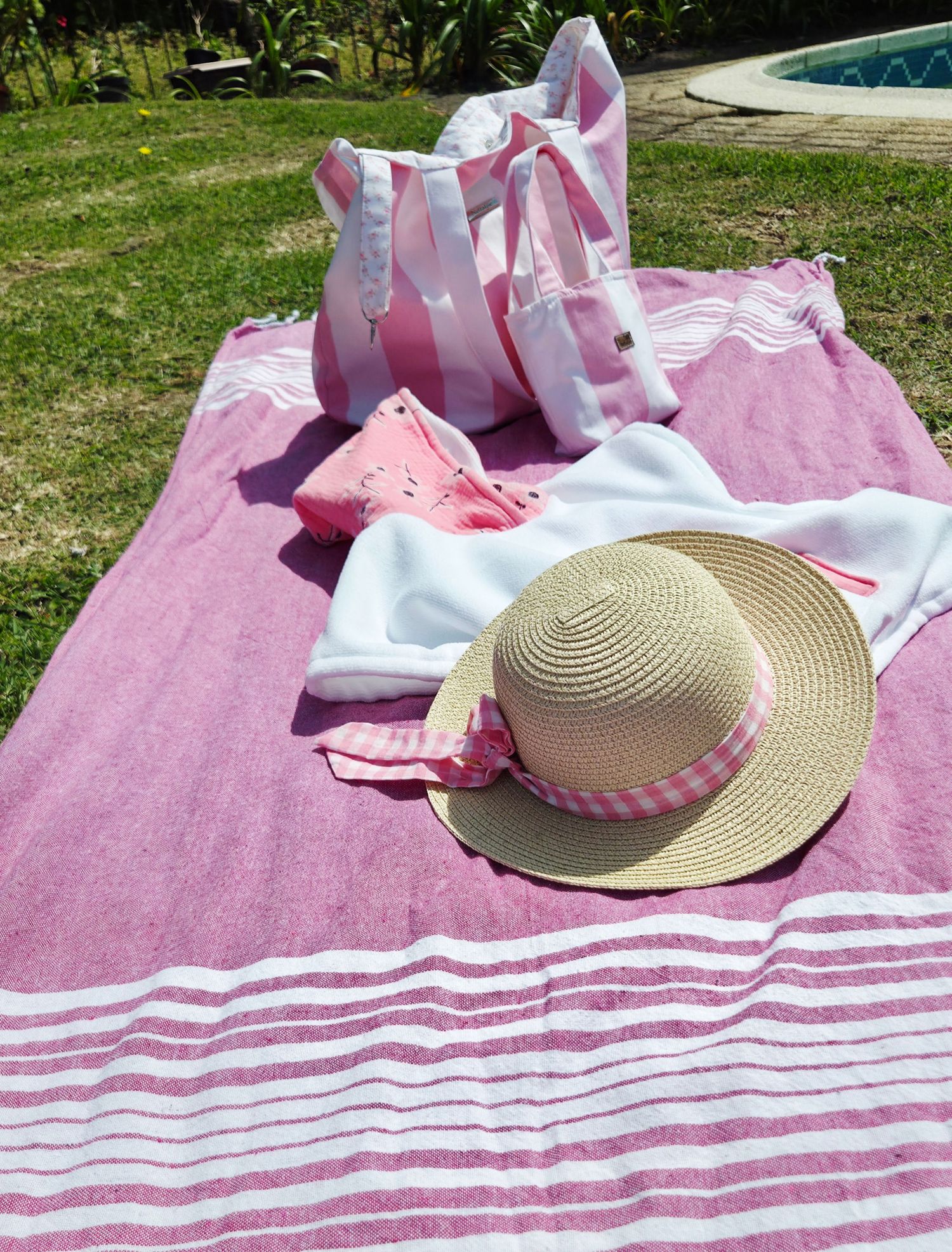 Pink striped beach towel with straw hat, pink gingham ribbon, tote bag, and accessories laid out on grass near pool.