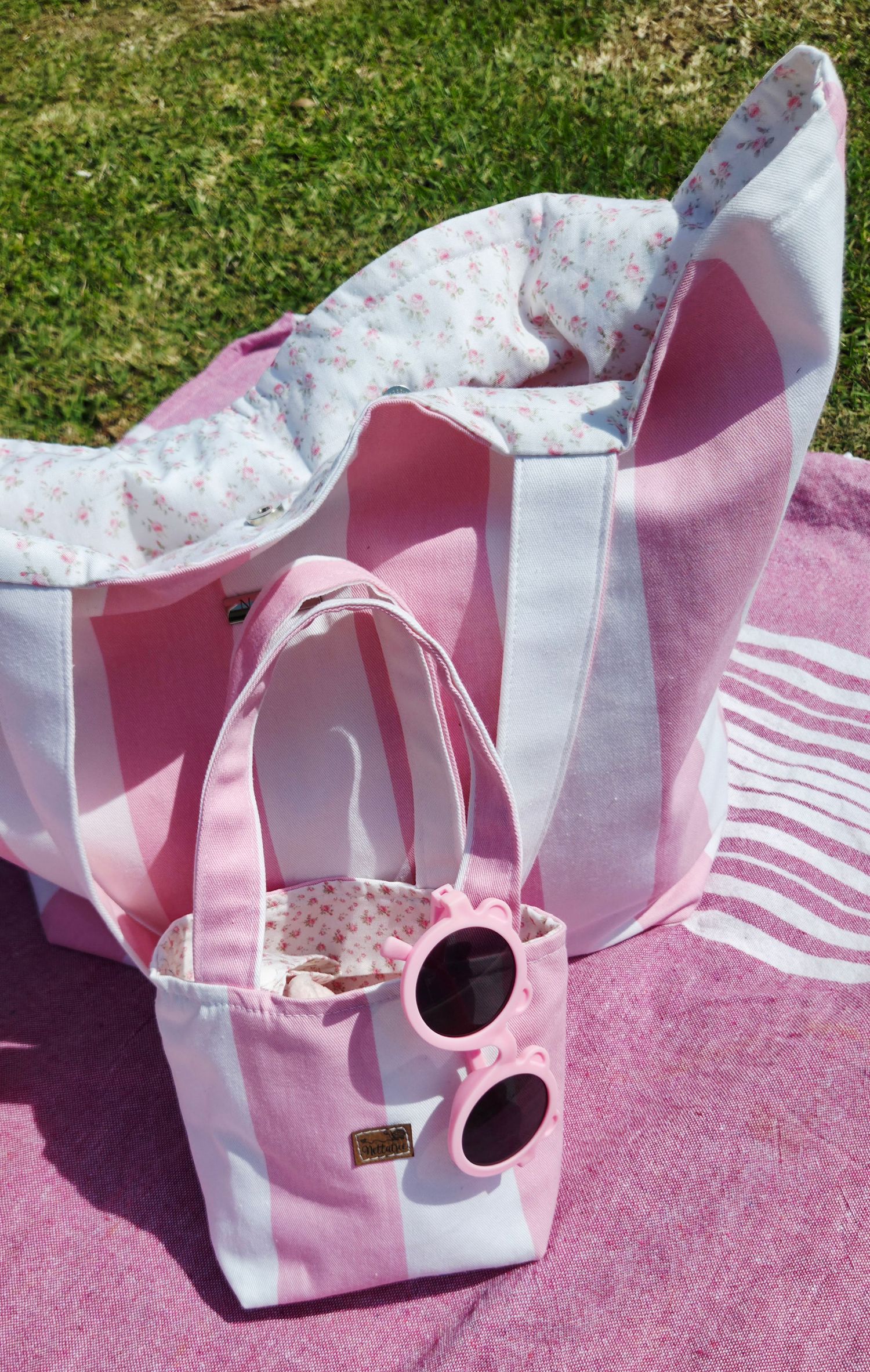 Pink striped beach tote bag with floral lining, mini matching bag, and pink sunglasses on a pink striped towel outdoors.