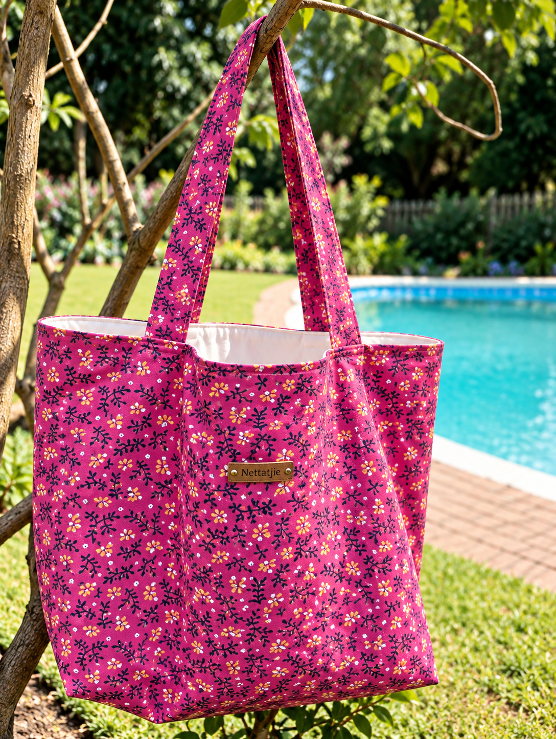 Pink floral tote bag with small colorful flowers hanging on a tree branch near a pool in a lush garden.