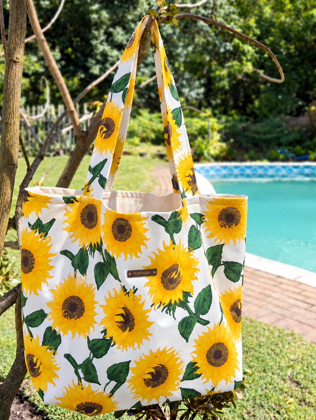 White tote bag with vibrant yellow sunflower print hanging on a branch near a pool in a lush garden setting.