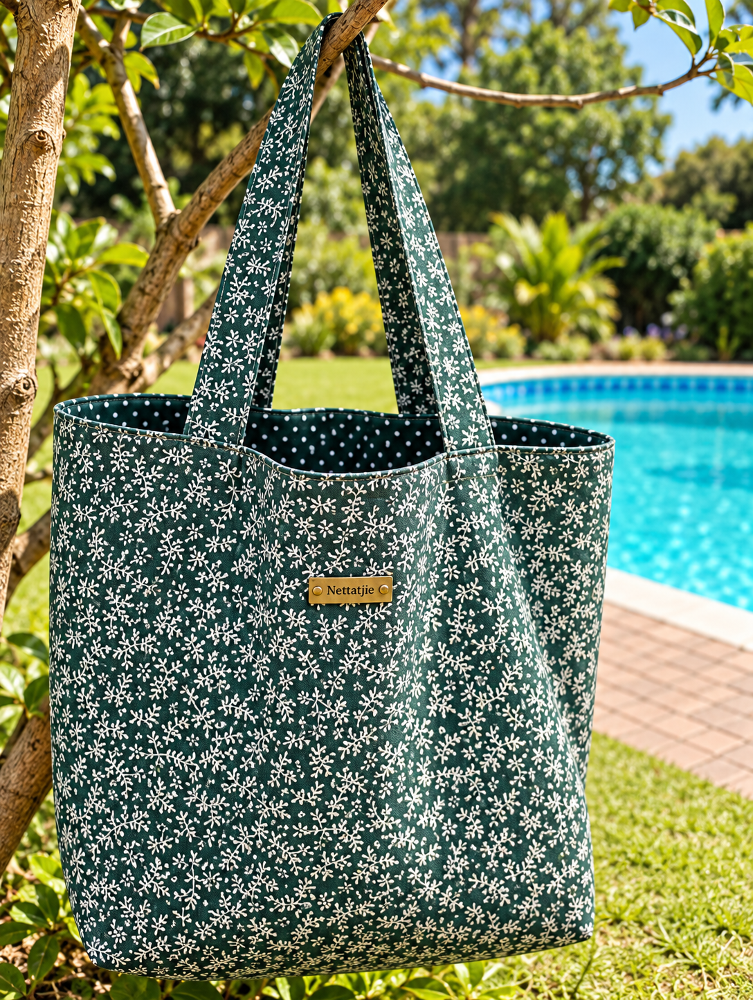 Green floral tote bag with white daisy print hanging on a tree branch near a bright blue swimming pool.