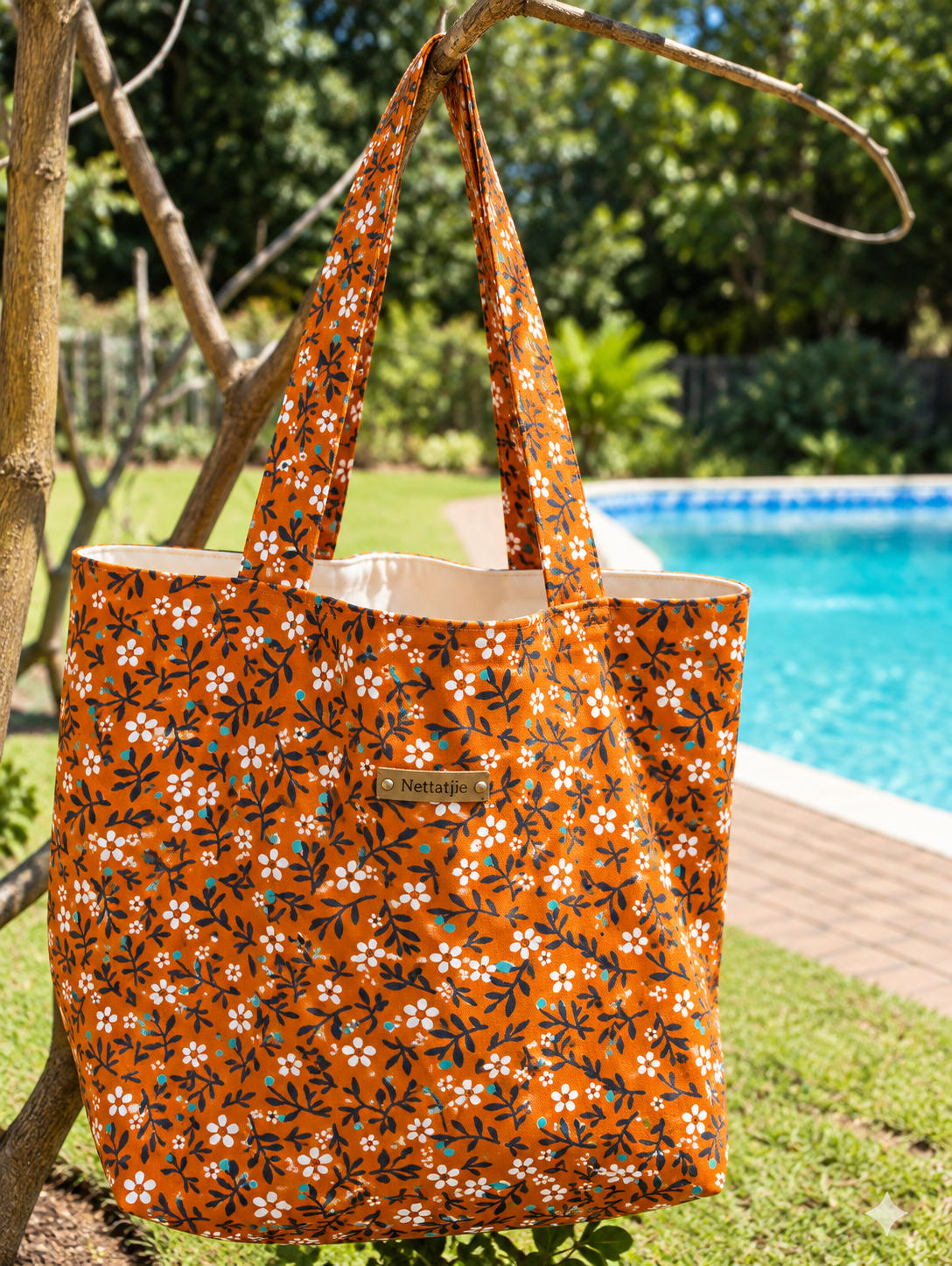 Orange floral tote bag with white daisy print hanging on a branch near a swimming pool on a sunny day.
