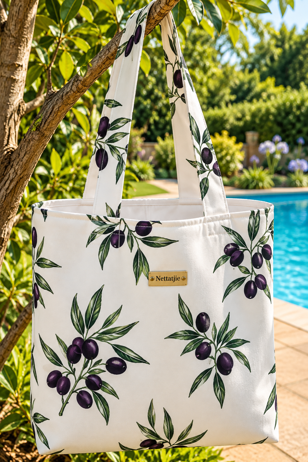 White tote bag with olive branch print pattern hanging on tree near pool, featuring Nettatjie brand label.