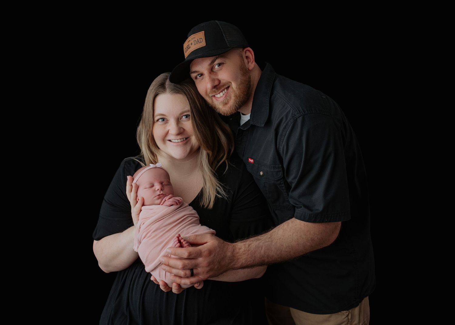 Happy couple in black outfits smiling while holding their newborn baby wrapped in pink against a dark background.