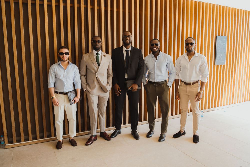 Five well-dressed men in smart casual and formal attire posing together in front of a wooden panel wall.