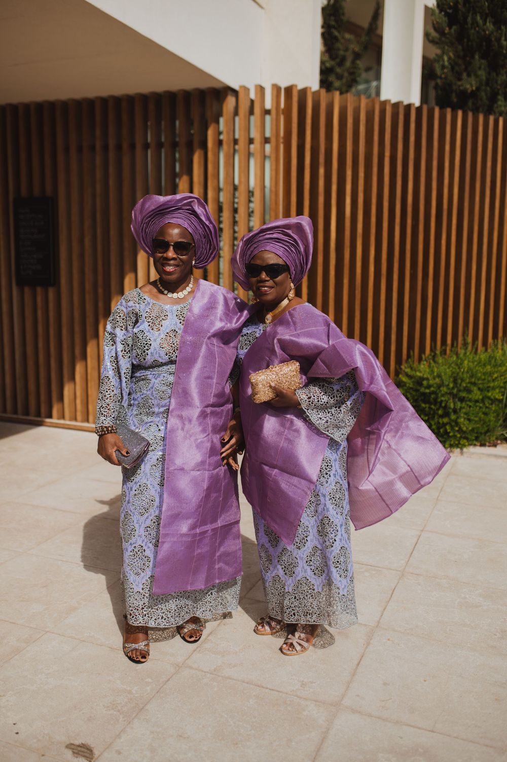 Two women in matching purple Nigerian traditional attire with gele head wraps and sunglasses posing outdoors.