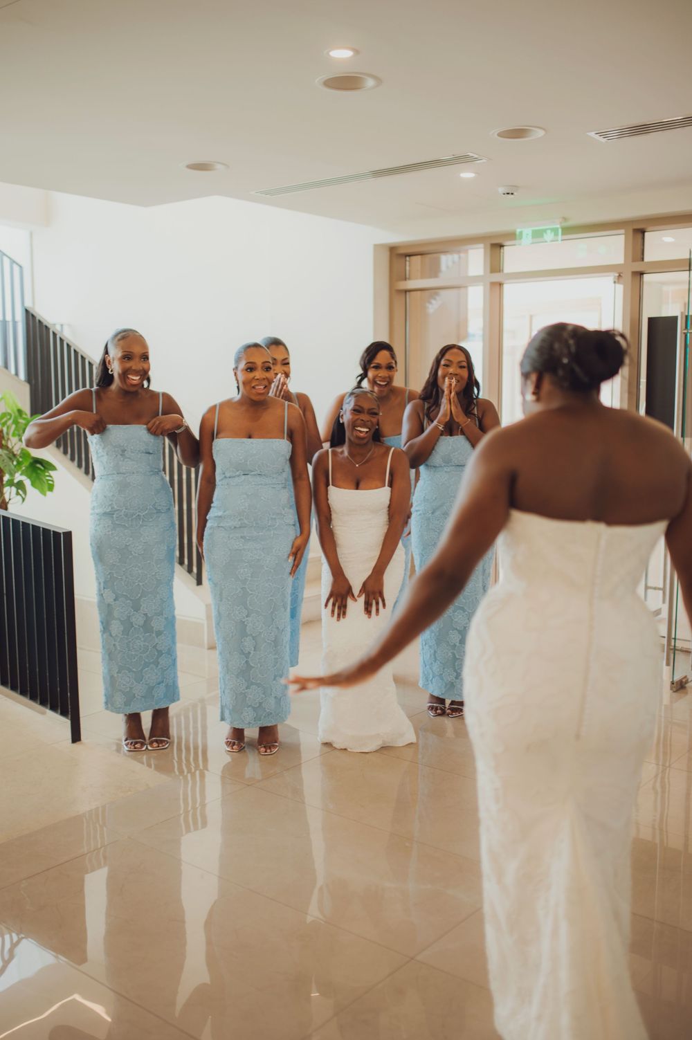 Bridesmaids in light blue lace dresses react with joy seeing the bride in her white wedding gown indoors.